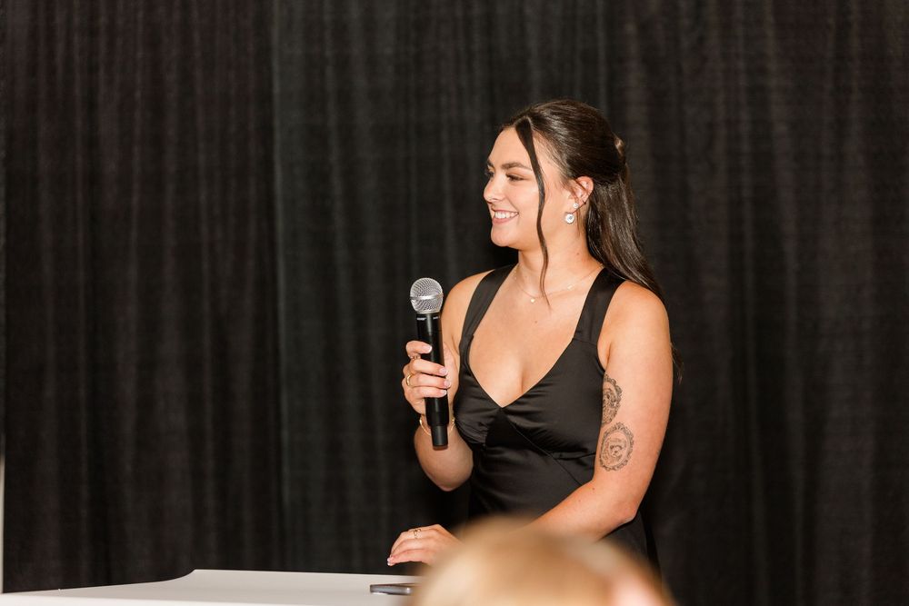 Woman in black dress holding microphone speaking at an event with dark curtain backdrop.