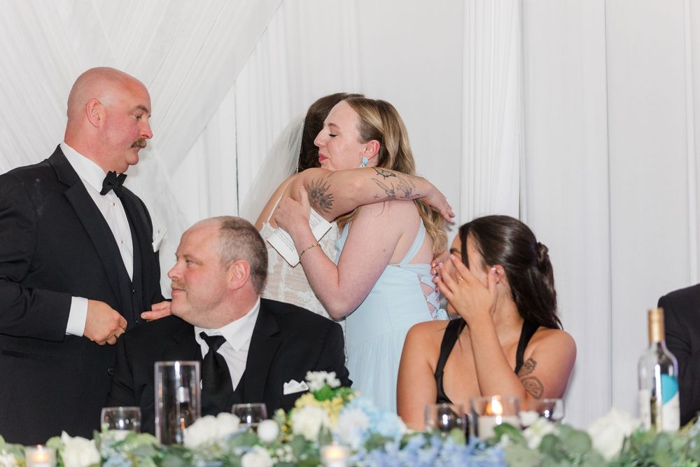 Two women share a warm hug at a wedding reception while guests in formal attire look on and smile.