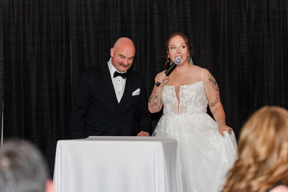 Bride in lace gown speaks into microphone beside groom in tuxedo at wedding reception with black drape backdrop.