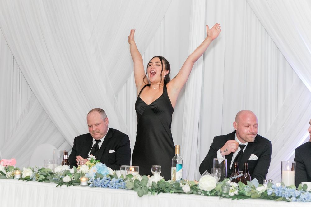 Woman in black dress raises arms joyfully while giving wedding speech at elegantly decorated head table.