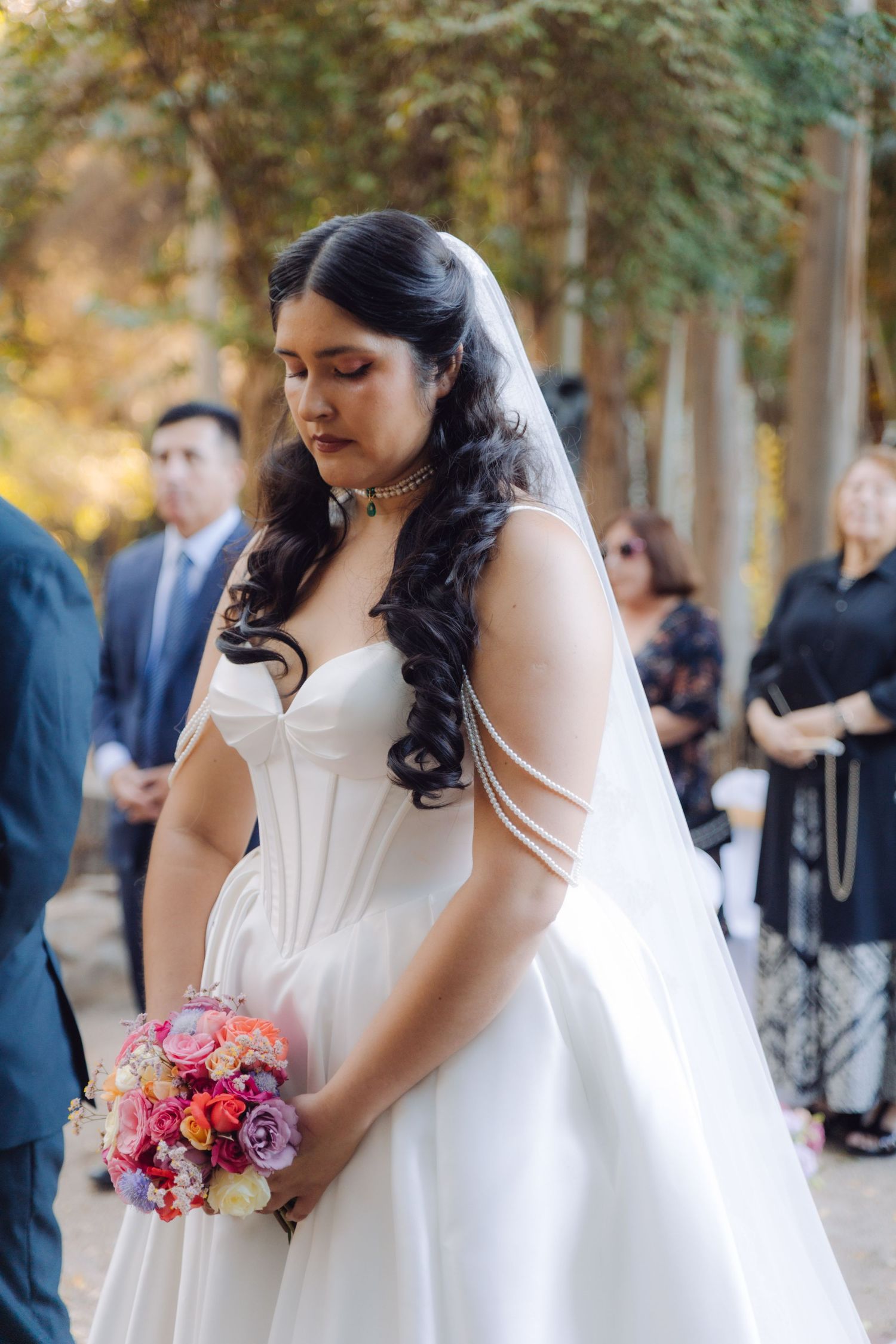 Beautiful bride in white corset wedding gown holding a colorful bouquet at an outdoor ceremony.