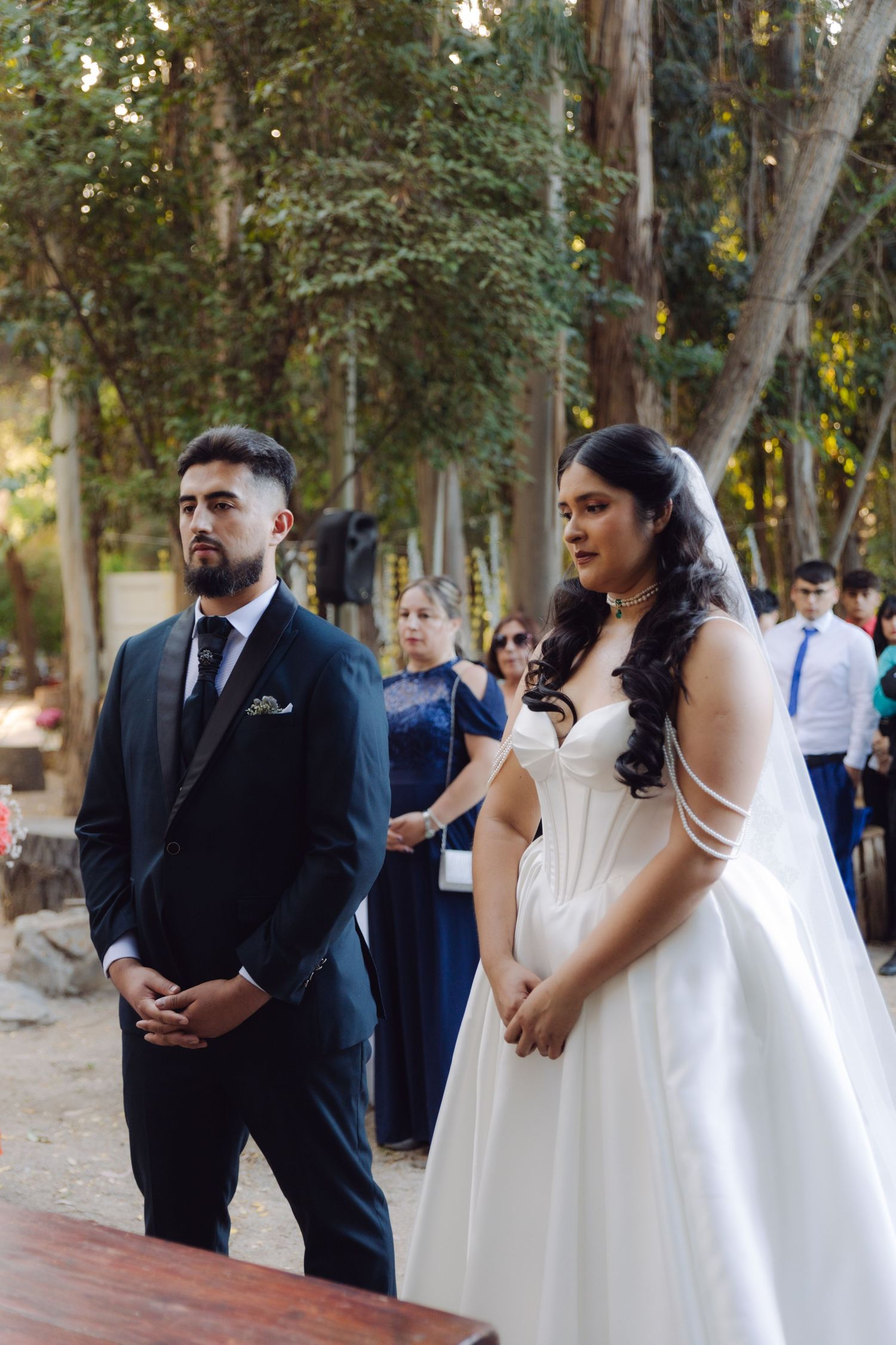 Bride and groom stand together attentively during their outdoor woodland wedding ceremony.