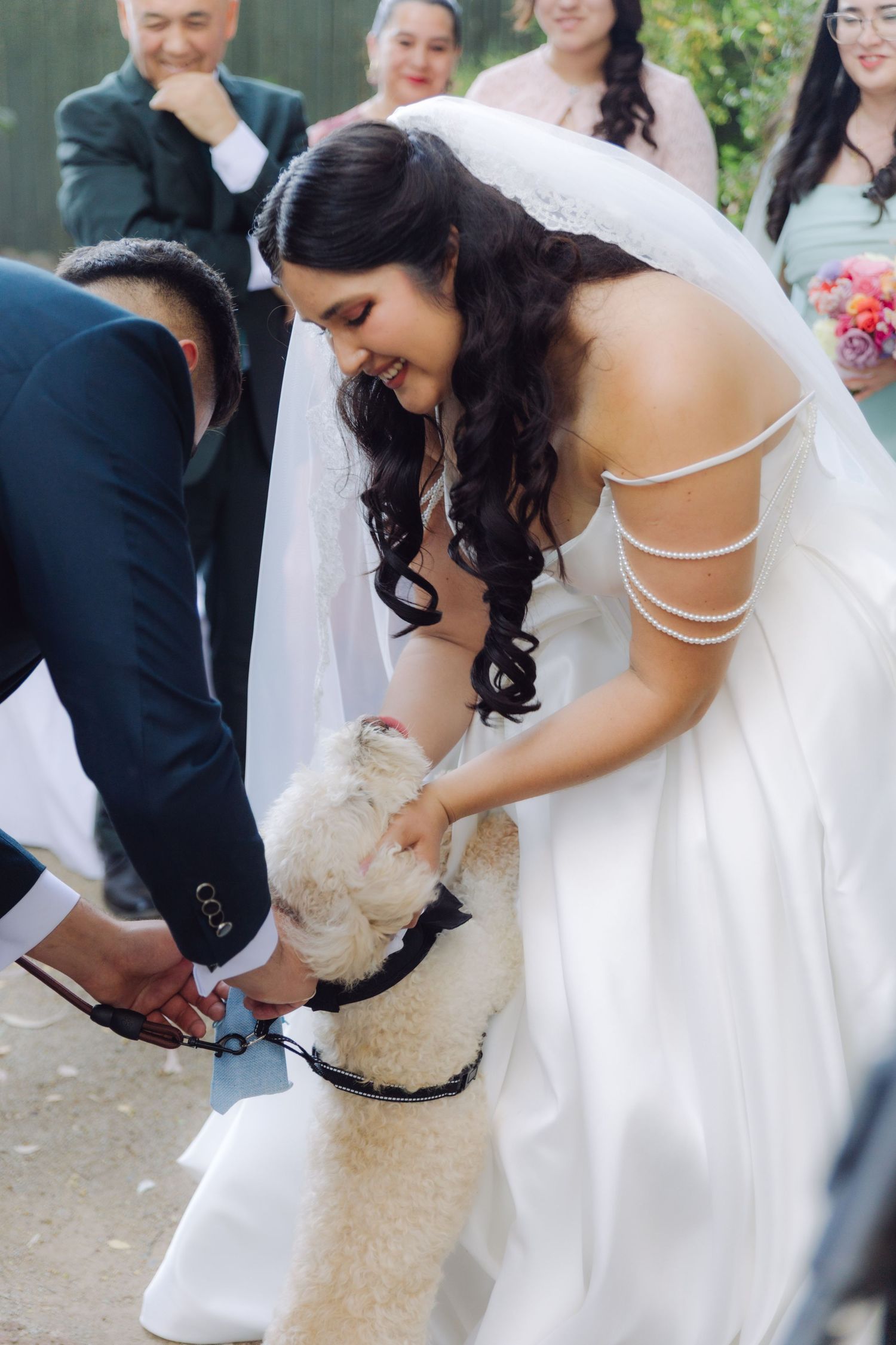 Smiling bride in white off-shoulder gown petting a fluffy cream dog during outdoor wedding ceremony.