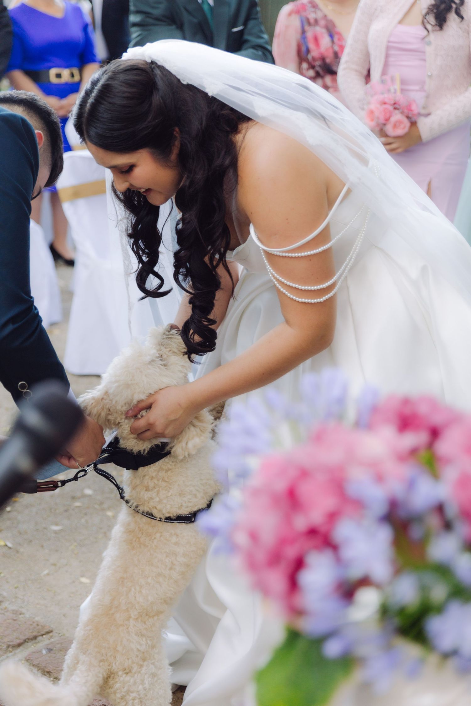 Bride in white wedding dress bending down to pet a fluffy cream-colored dog at outdoor ceremony.