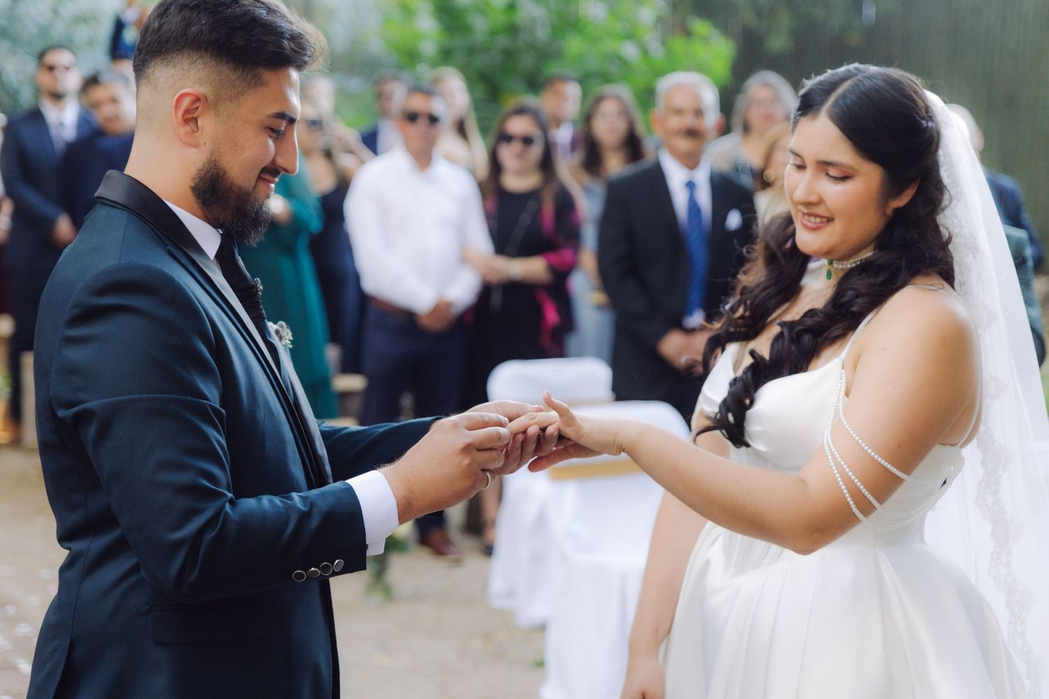 Groom in navy suit placing wedding ring on brides finger during outdoor ceremony with guests.