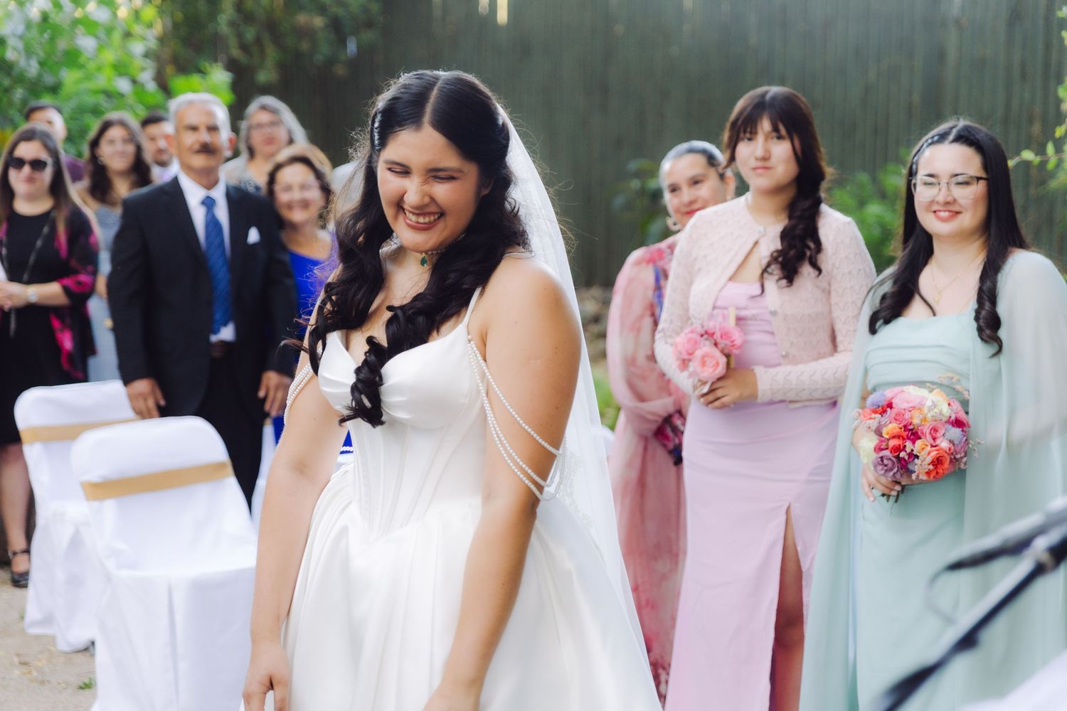 Bride in white gown and veil walking down aisle with bridesmaids in pink and sage green dresses.