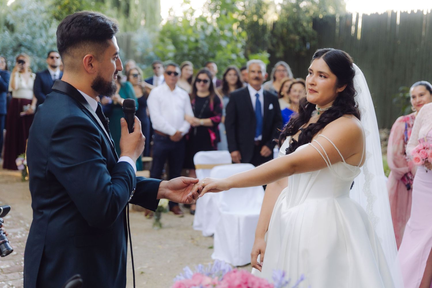 Bride and groom holding hands during outdoor wedding vows with guests gathered in background.