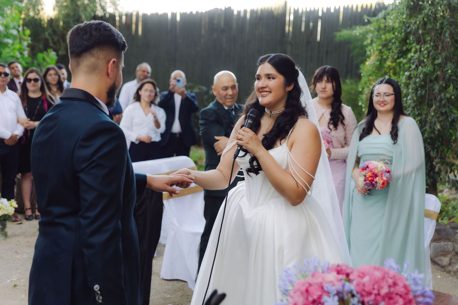 Smiling bride in white gown speaking vows into microphone while groom holds her hand outdoors.