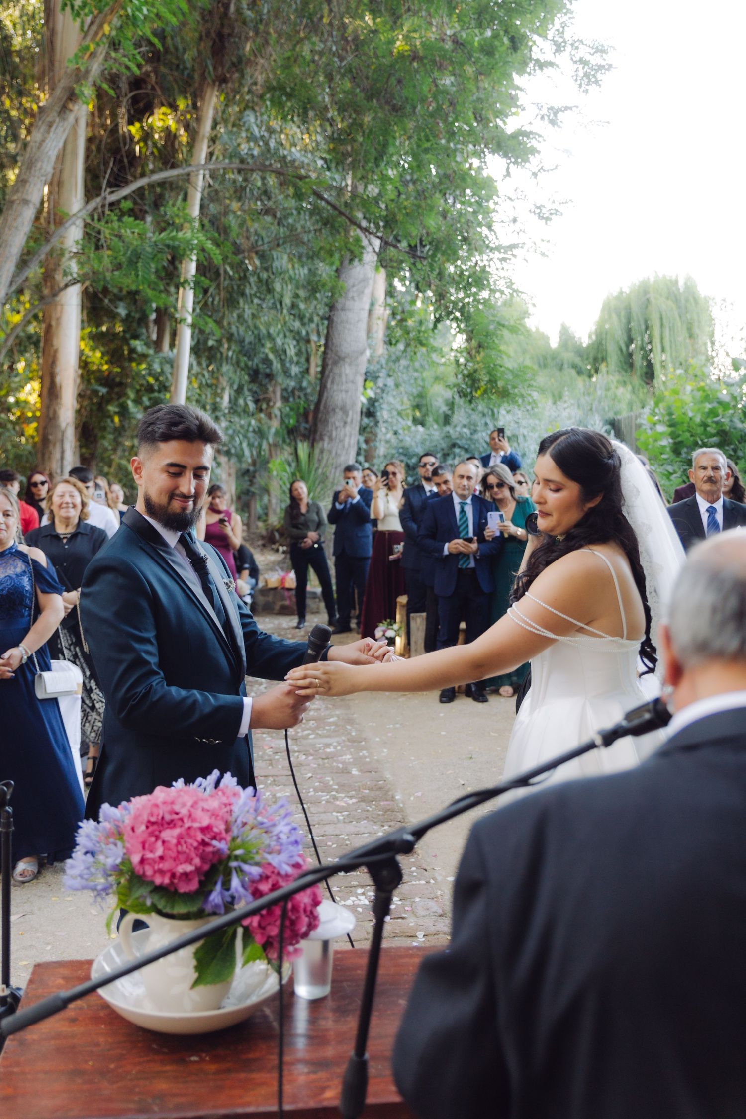 Bride and groom exchange rings during outdoor wedding ceremony with guests watching among lush green trees.