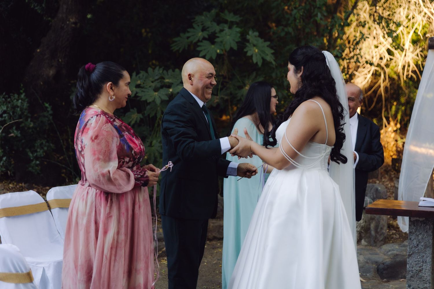 Bride in white gown greets smiling bald man and woman in pink dress at outdoor evening wedding reception.