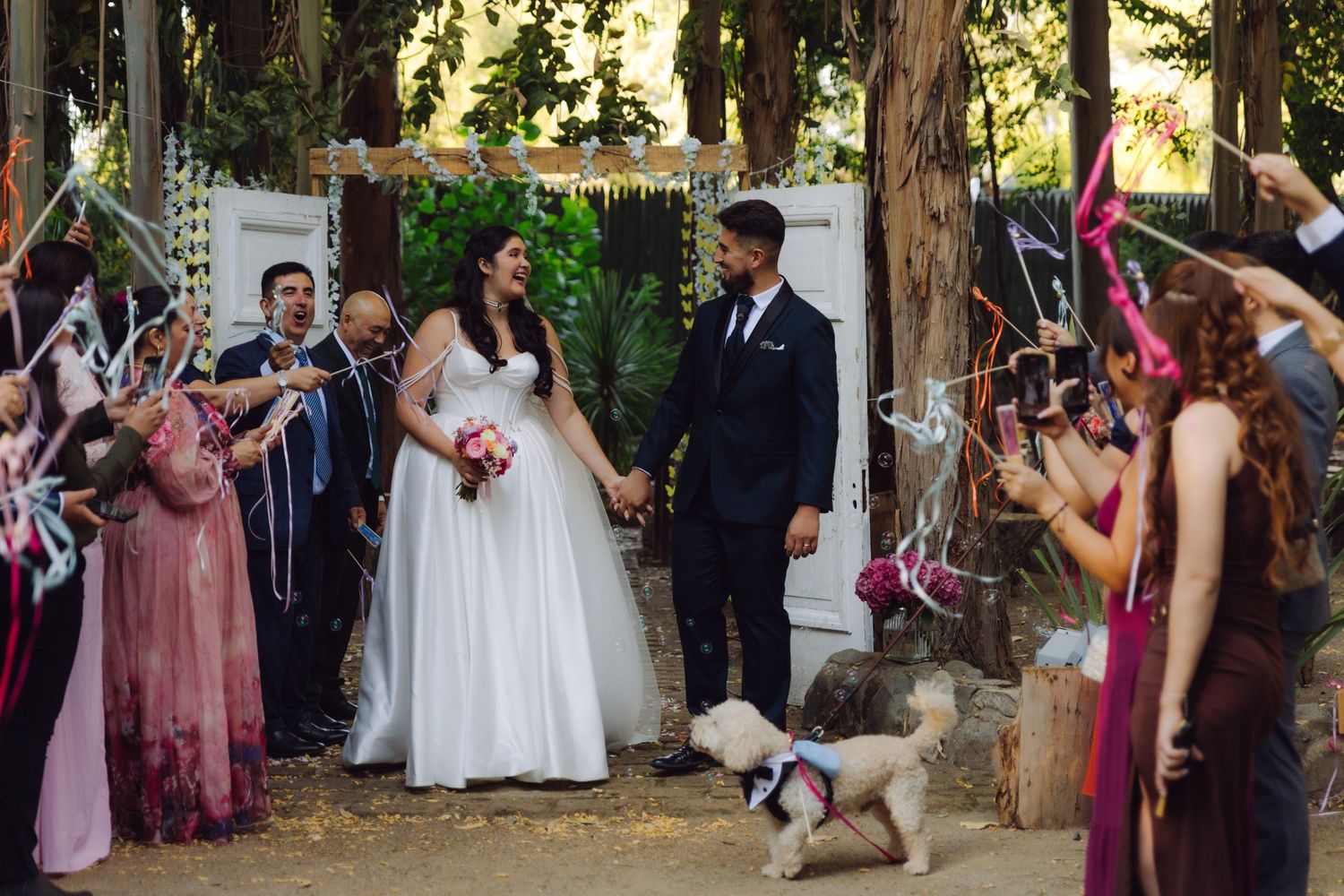 Newlyweds hold hands with small white dog at feet during ribbon wand exit at outdoor forest wedding.