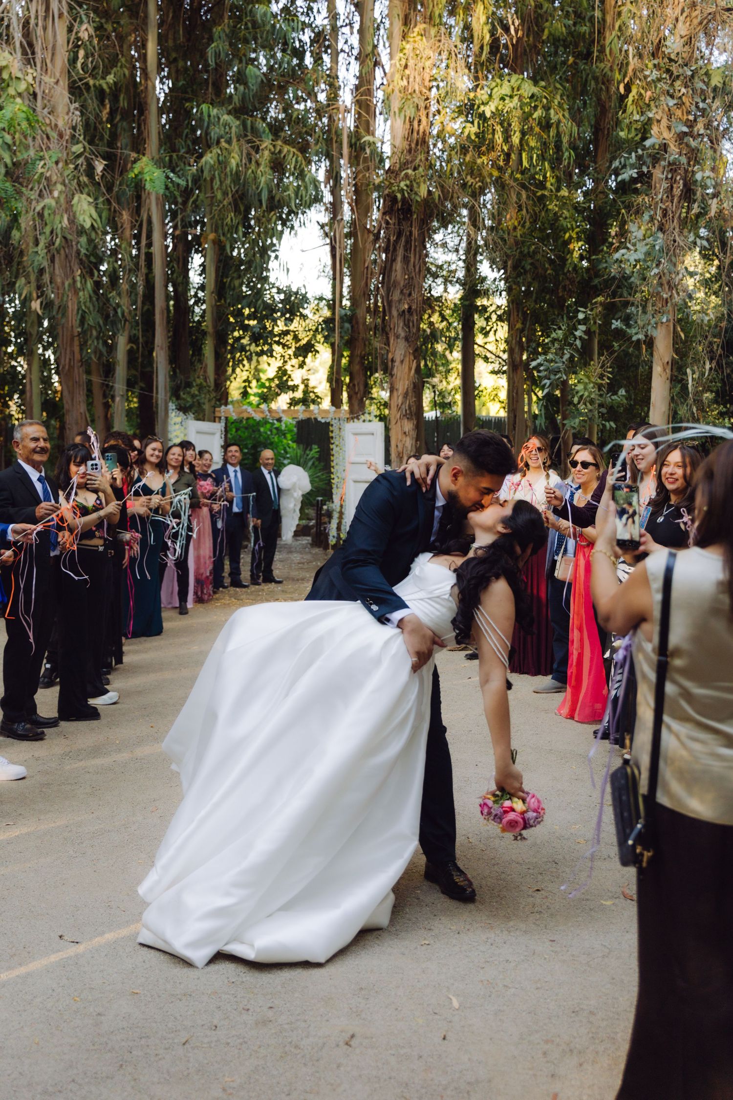 Groom dips bride for passionate kiss during outdoor wedding sendoff lined with cheering guests and ribbons.