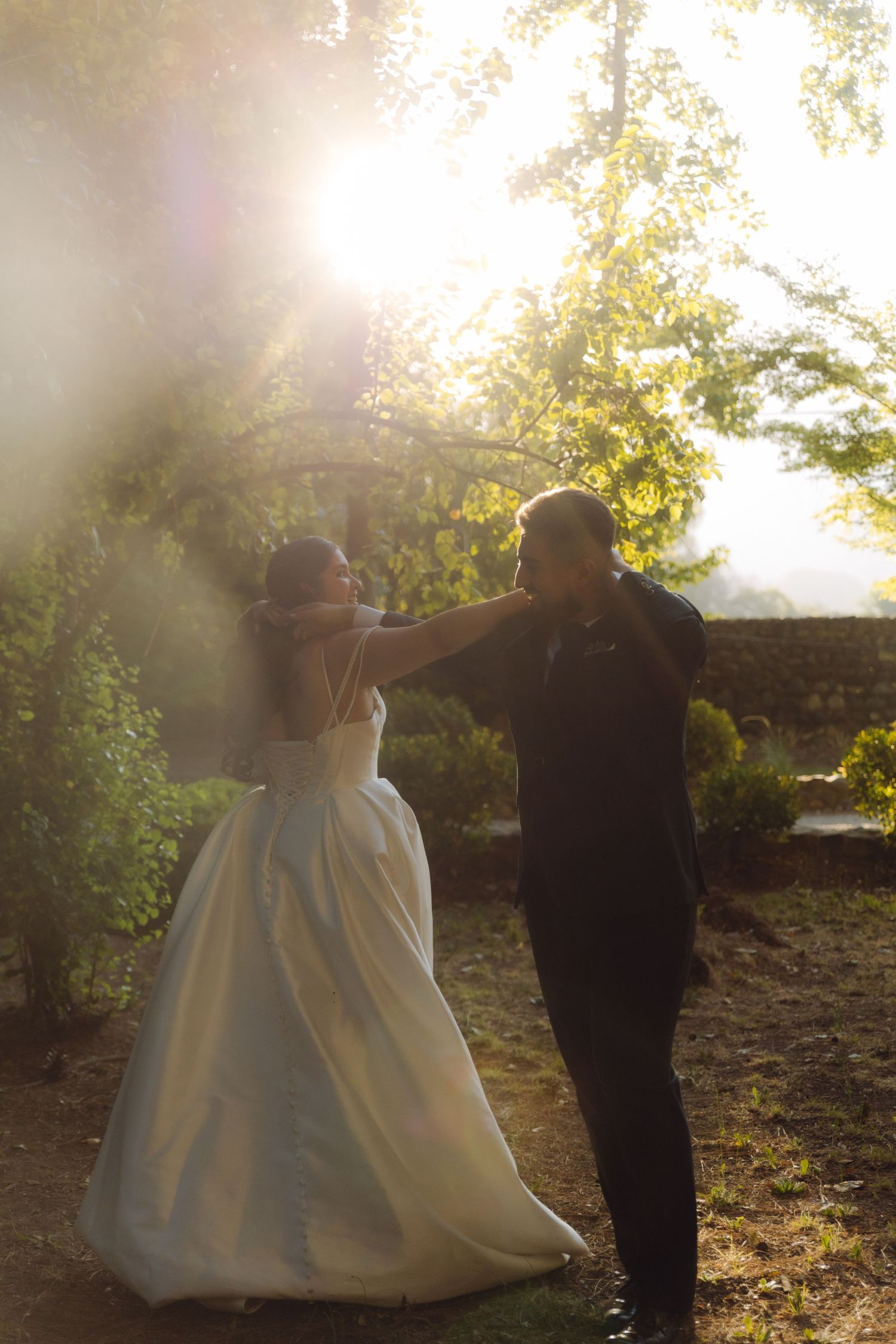 Bride and groom dancing in golden sunlight beneath trees during outdoor wedding shoot.