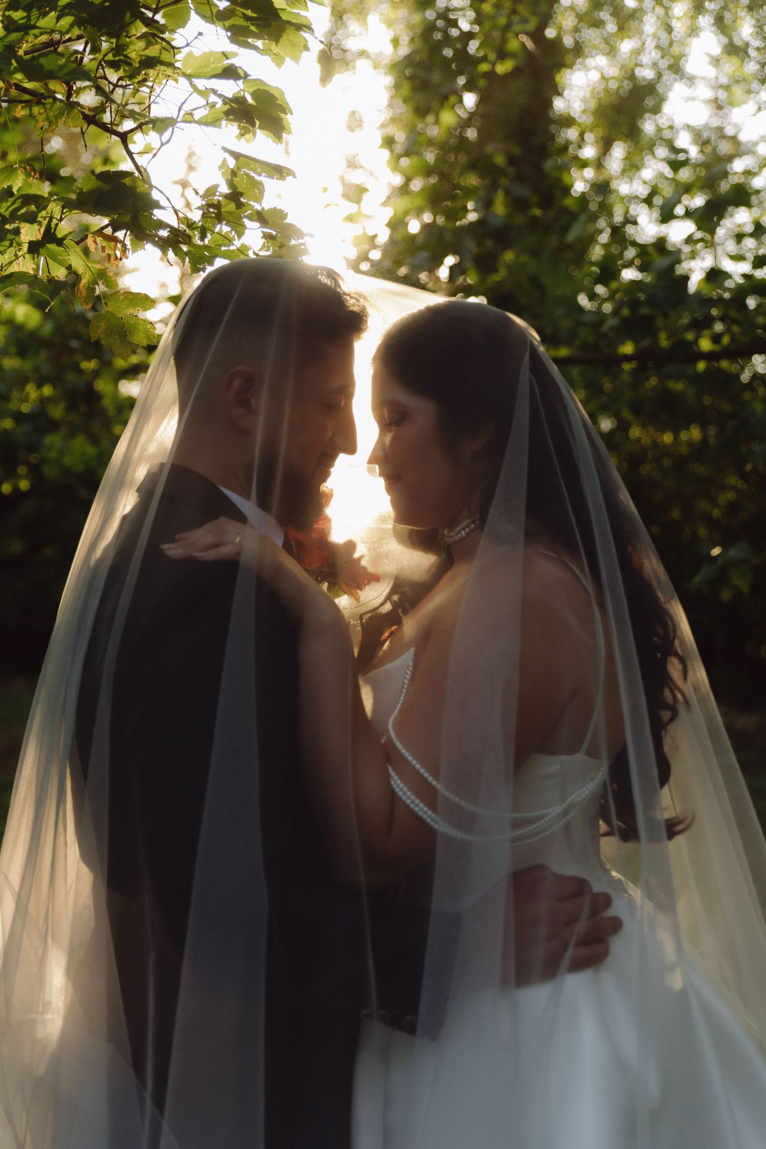 Bride and groom sharing intimate moment under veil in glowing sunset garden setting.