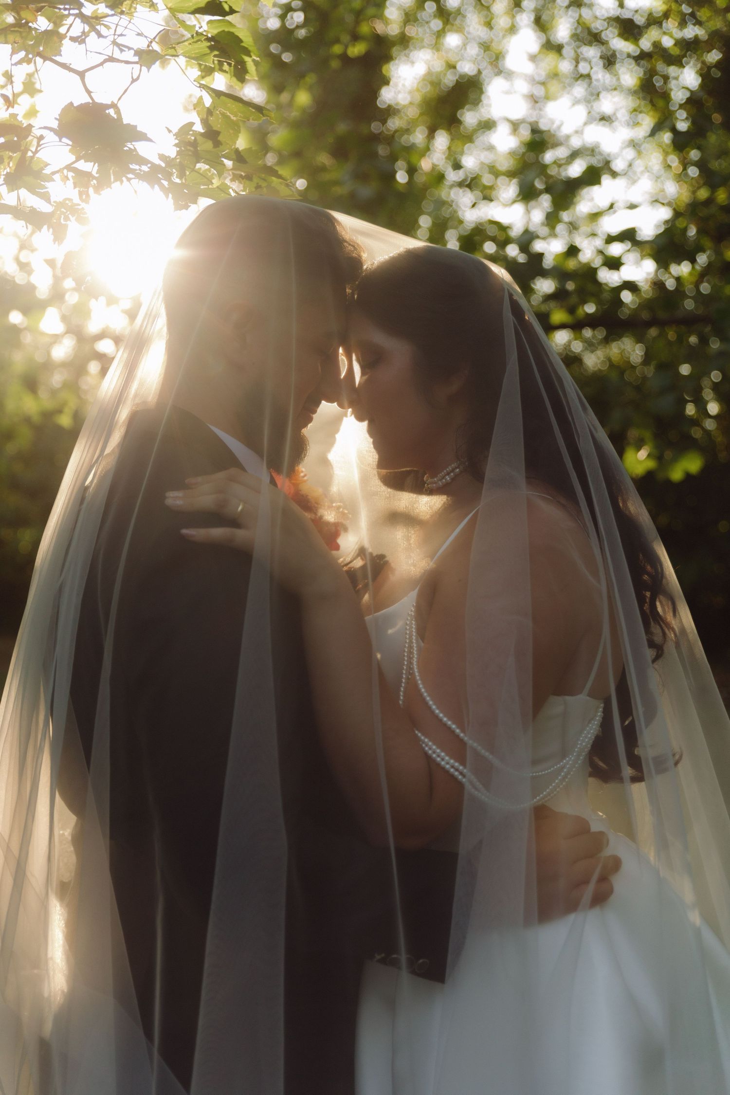 Bride and groom embracing under flowing veil in warm golden sunset garden light.