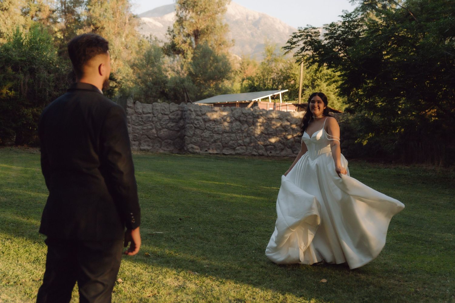 Bride in elegant white gown spins joyfully on grass while groom in black suit watches at sunset.