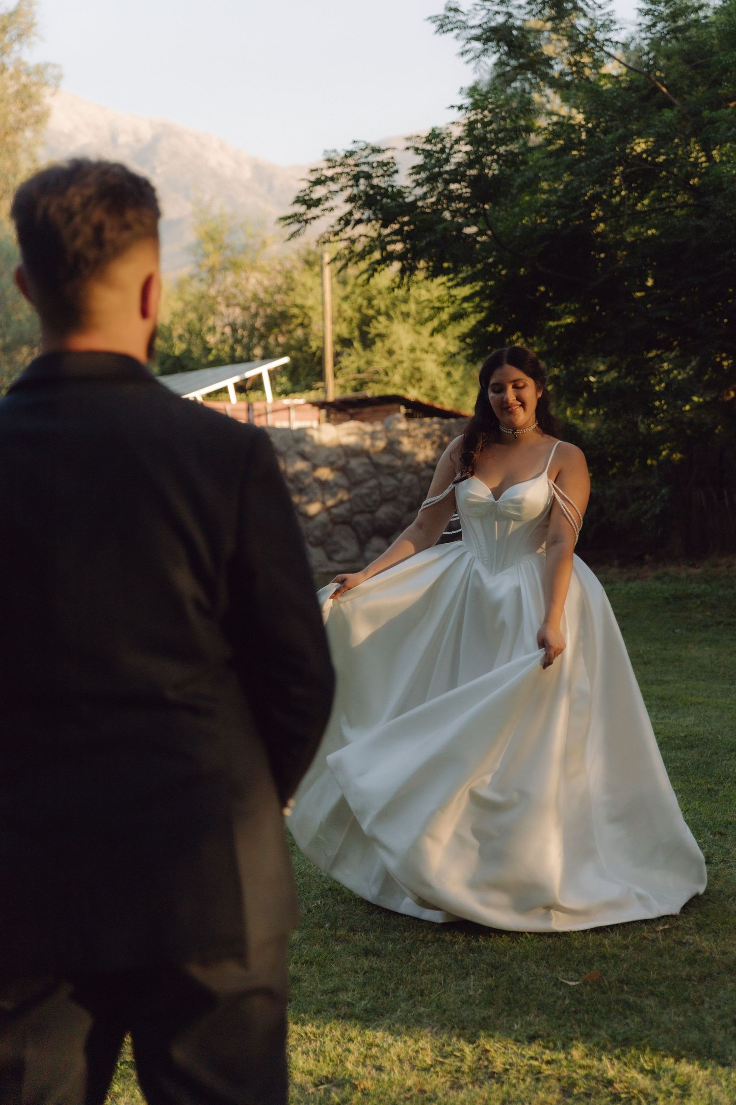 Smiling bride in a white satin ball gown twirls on green lawn as groom watches in golden sunset light.