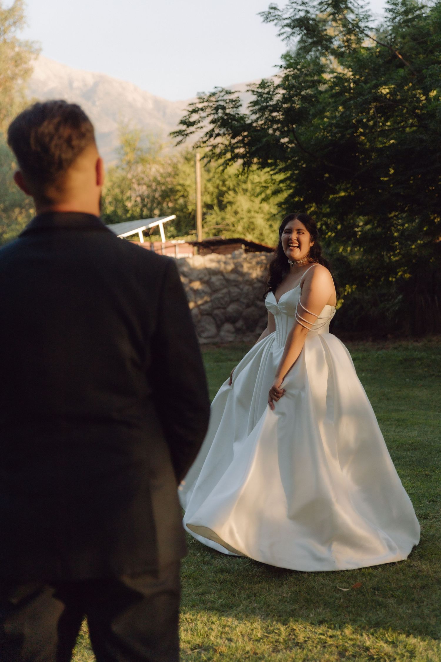 Laughing bride lifts her white satin wedding dress while approaching her groom on a sunlit garden lawn.