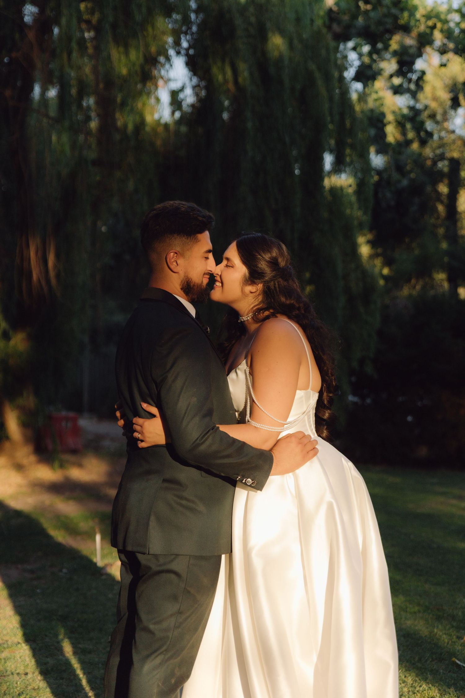 Bride and groom share a romantic kiss while embracing in a lush green garden at golden hour.