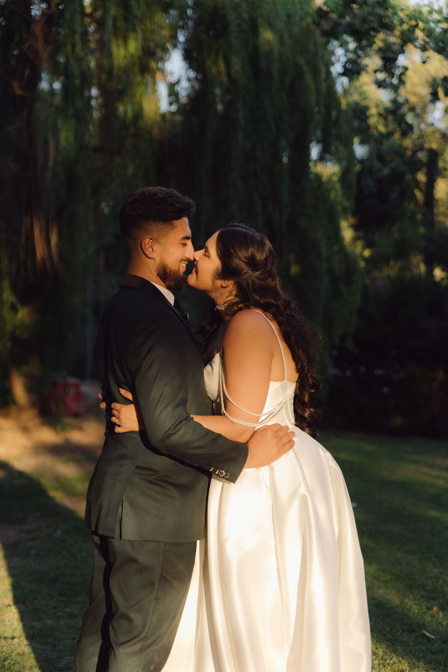 Newlyweds embrace and lean in for a kiss surrounded by tall green trees in warm sunset light.
