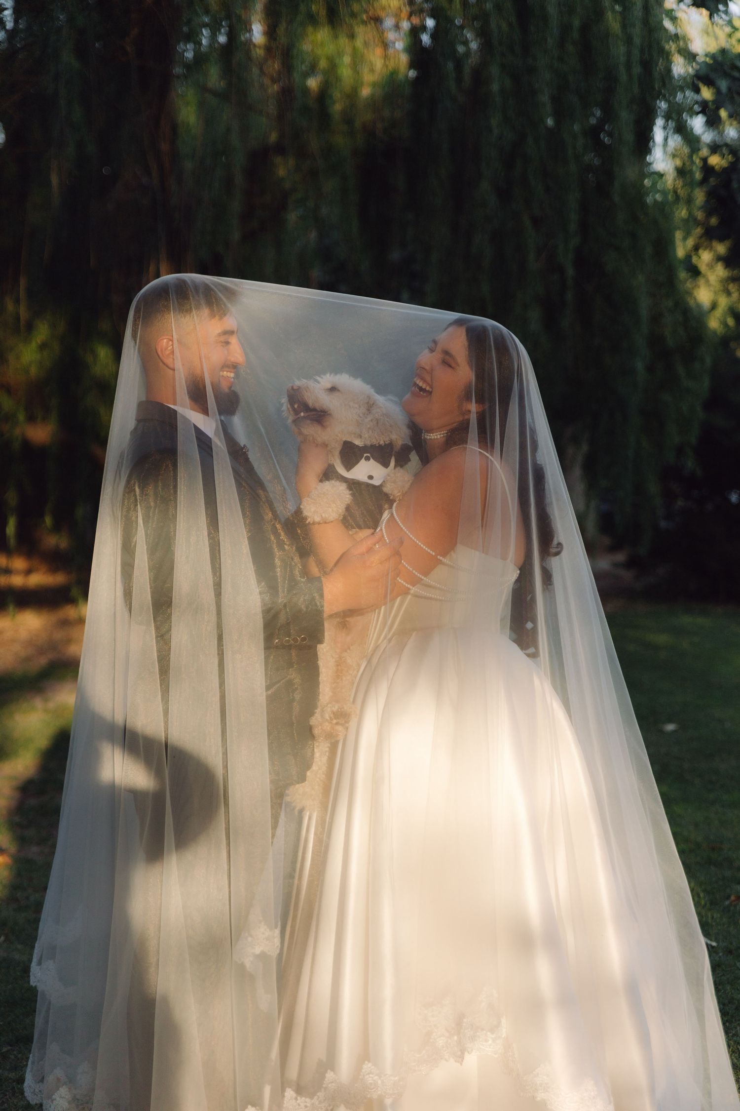 Laughing bride and groom hold their fluffy white dog under a wedding veil in a green garden.