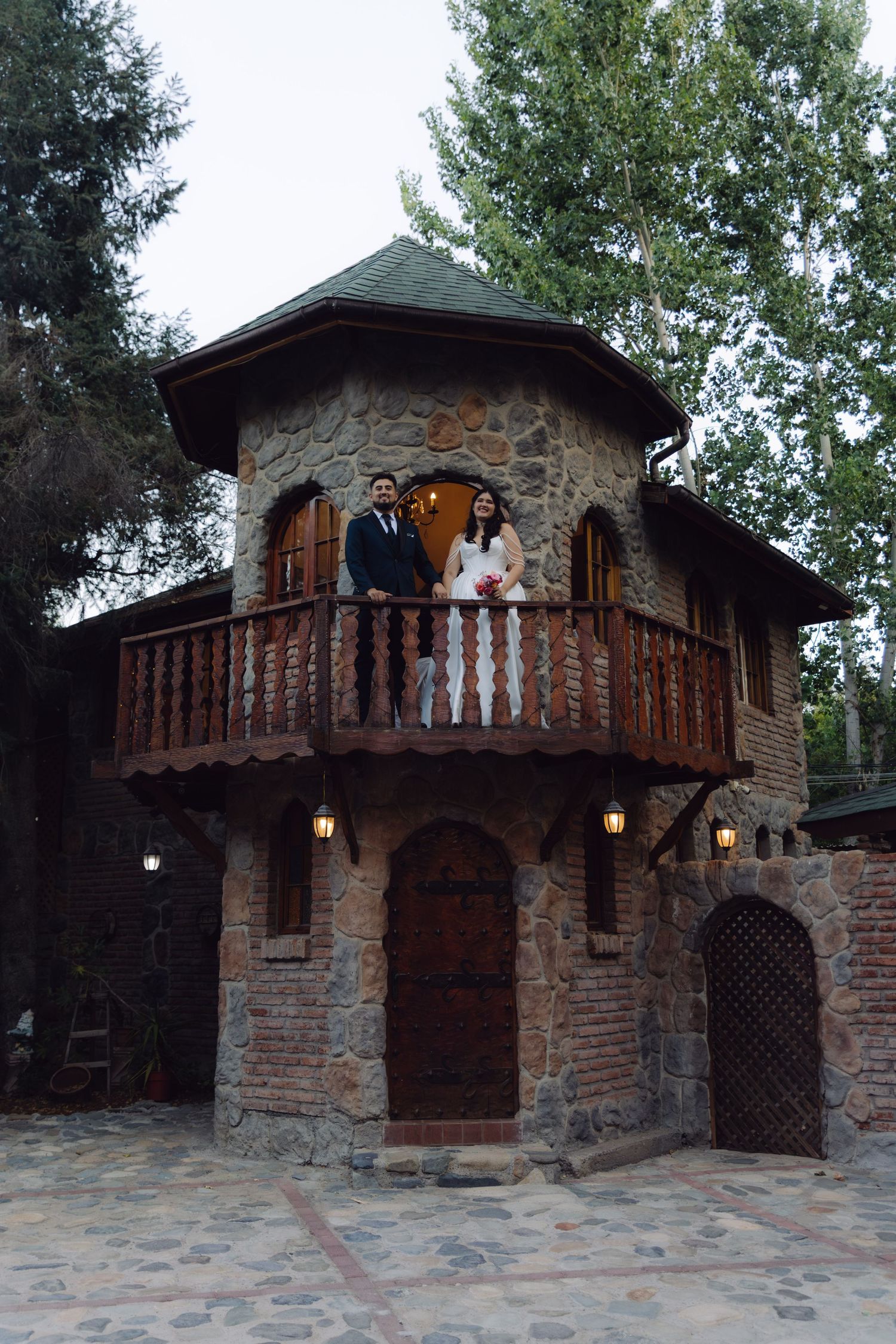 Wedding couple poses on the balcony of a charming stone castle tower venue surrounded by trees.