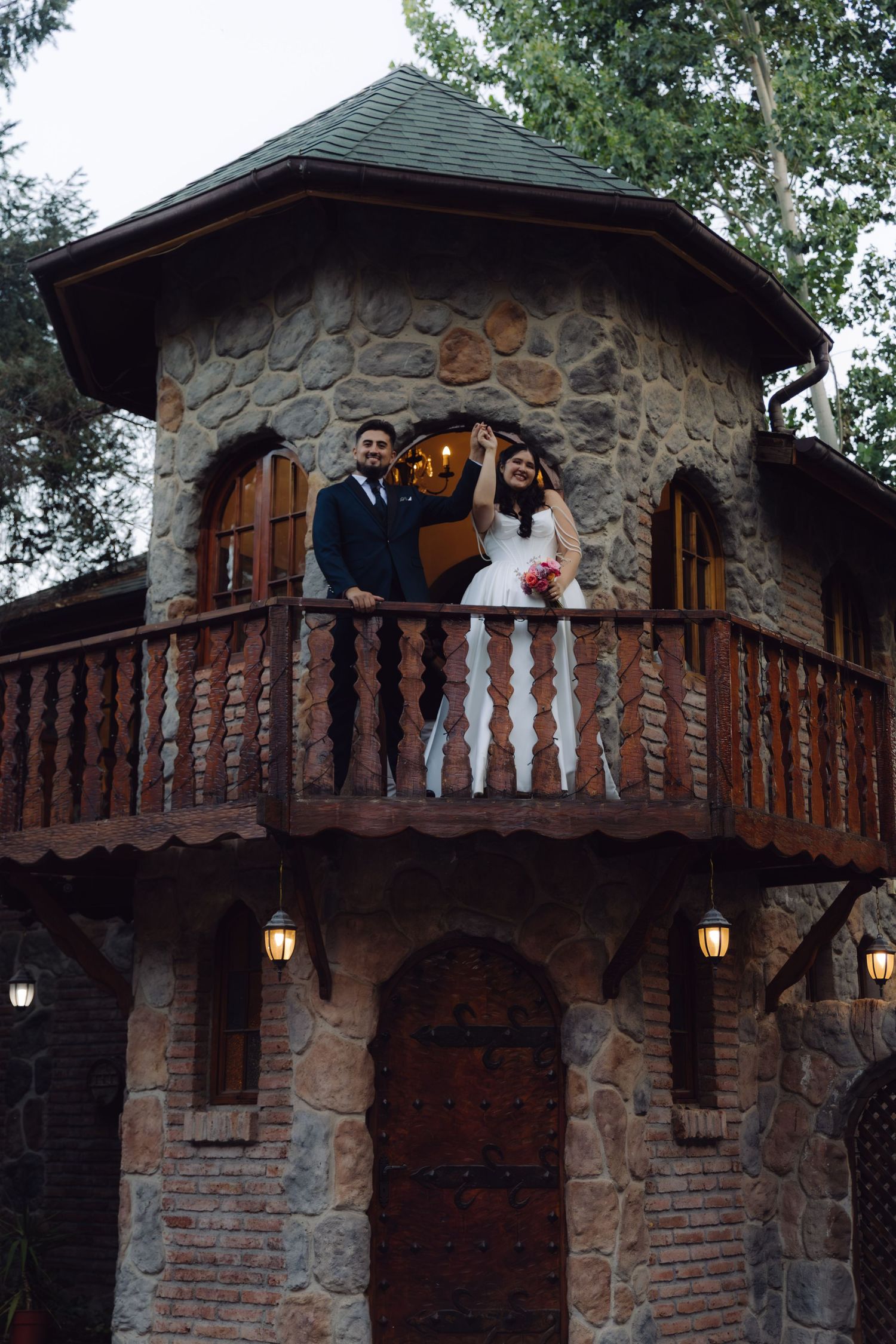 Joyful bride raises her hand while standing with groom on a rustic stone castle balcony at wedding.