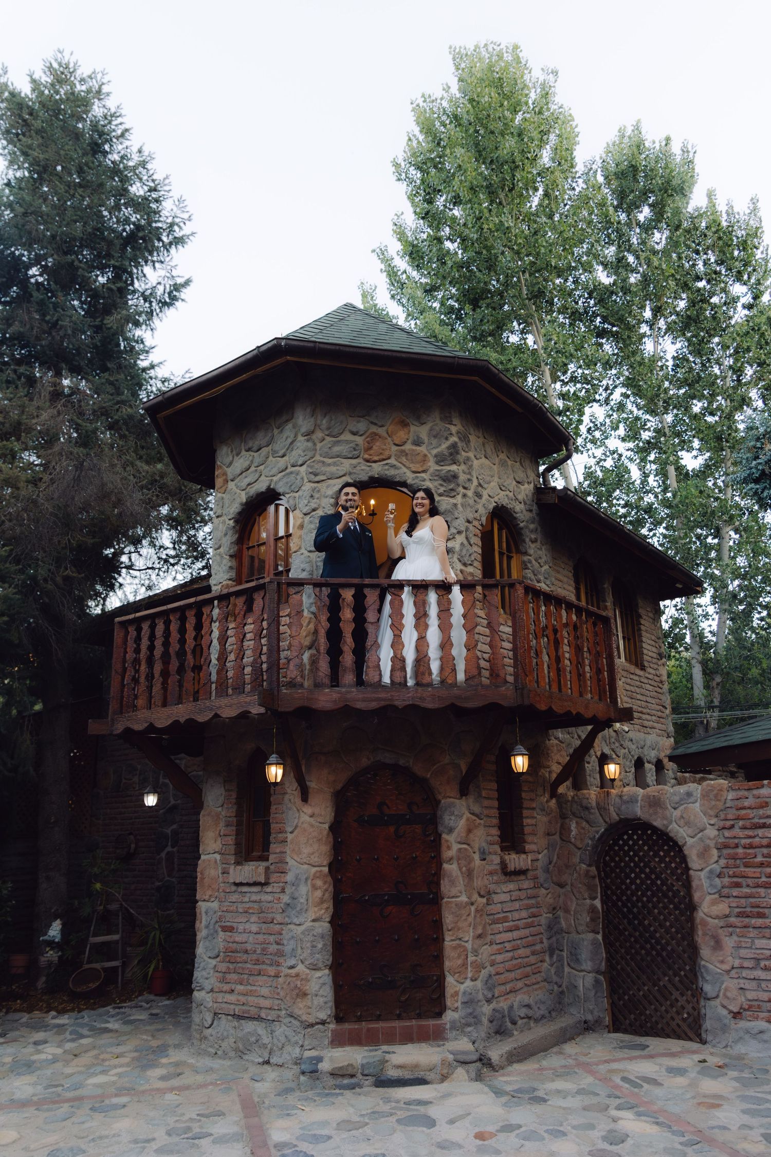 Wedding couple stands on the balcony of a picturesque stone tower castle venue at dusk.