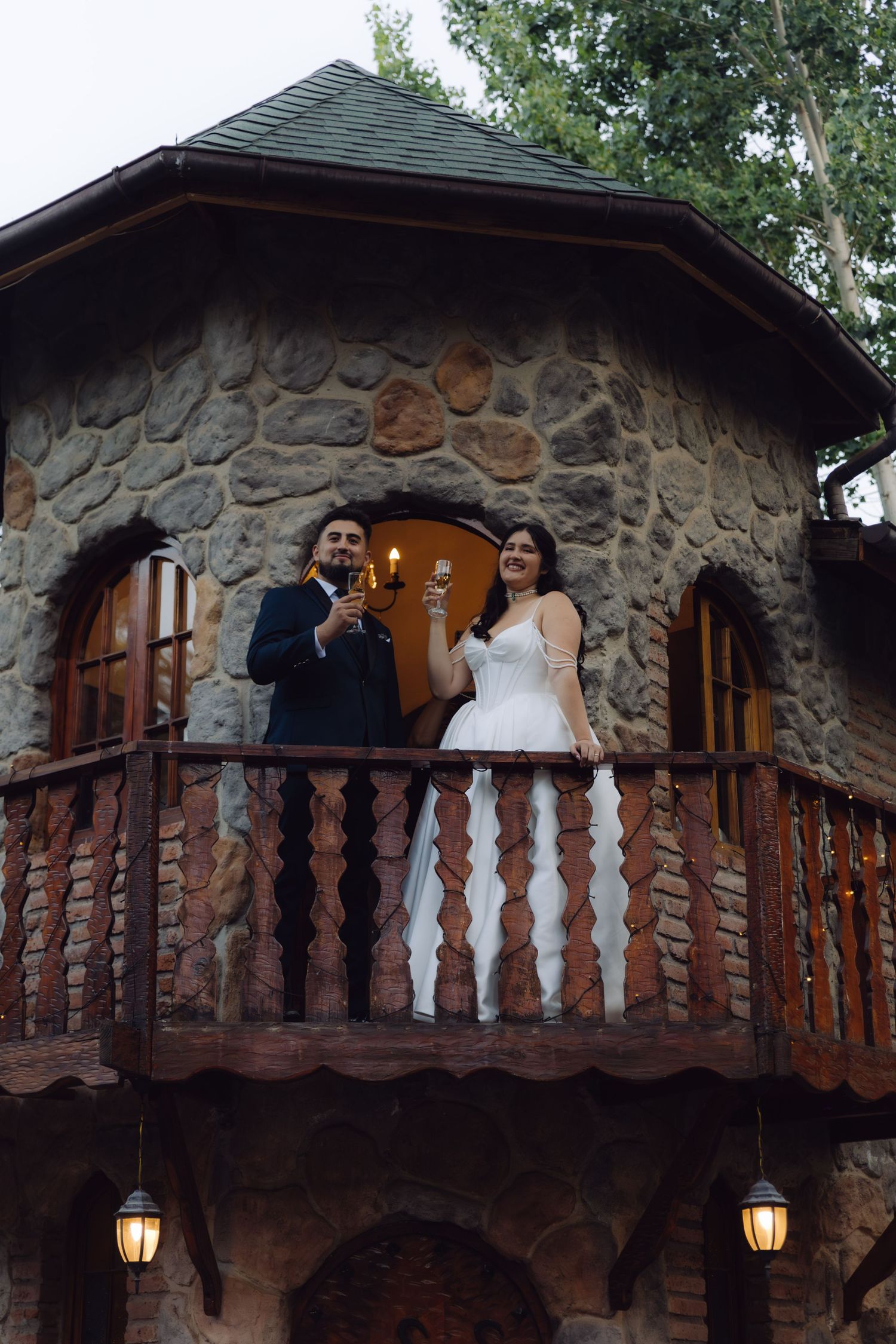 Smiling bride and groom raise champagne glasses in celebration on a rustic stone castle balcony.