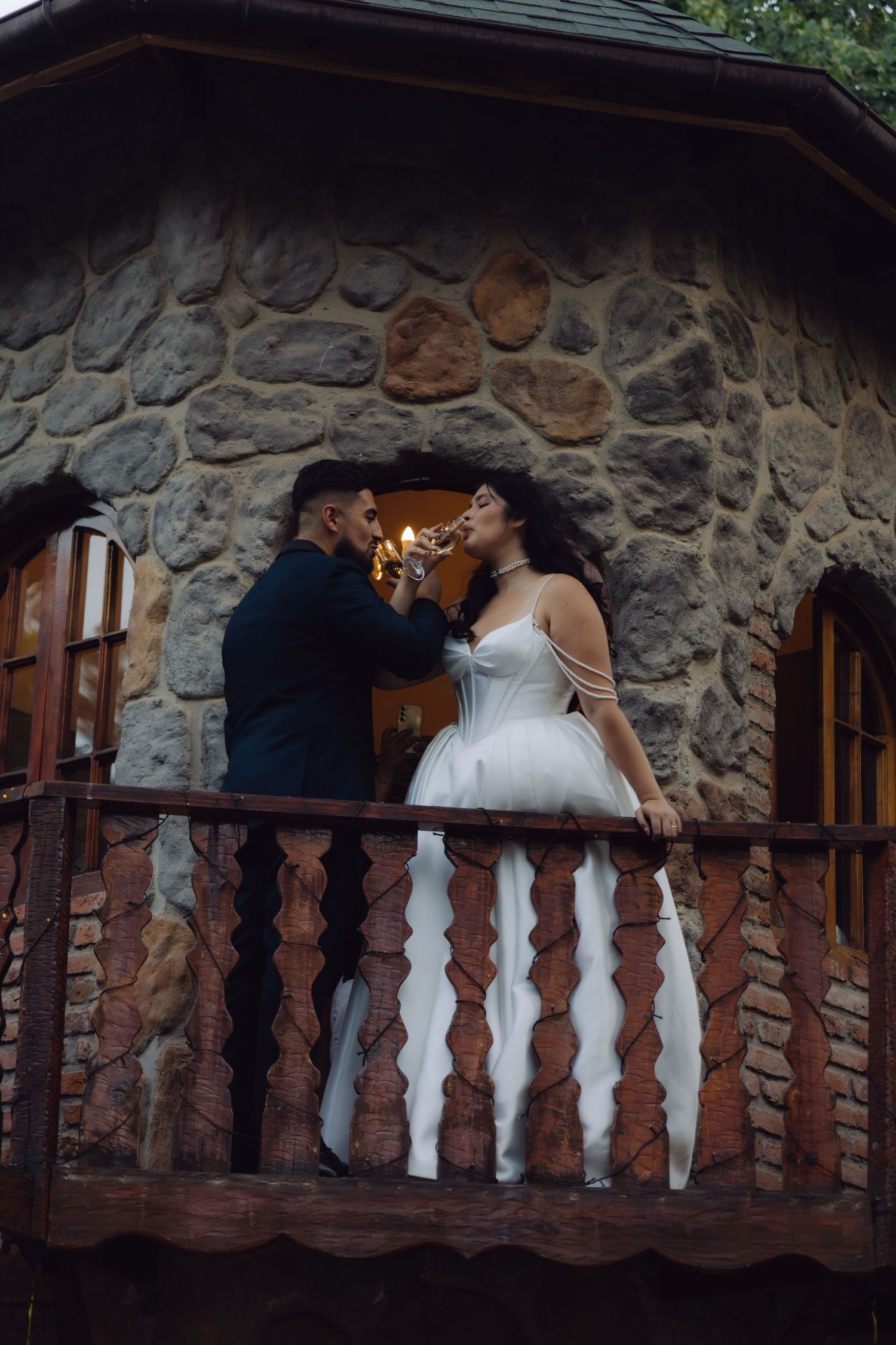 Groom offers champagne to bride on a wooden balcony of a stone castle wedding venue at dusk.