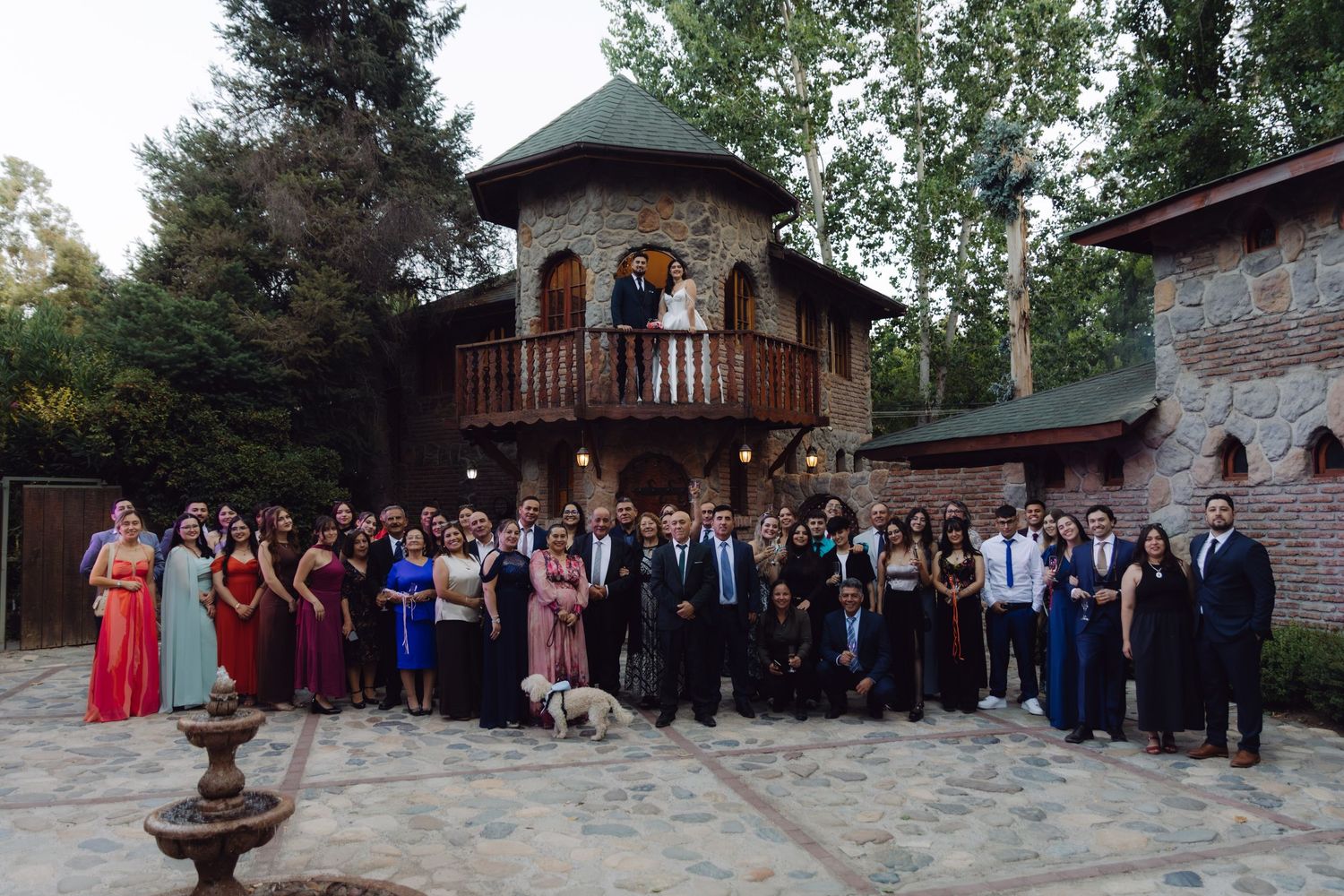 Large group of formally dressed wedding guests pose in front of stone castle venue.