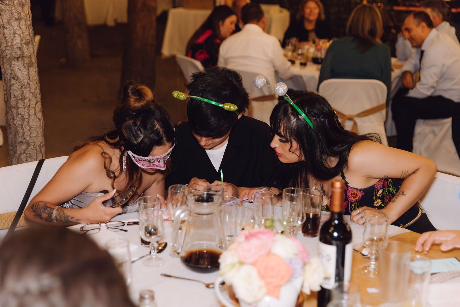 Wedding guests wearing festive headbands and masks leaning over table during reception celebration.