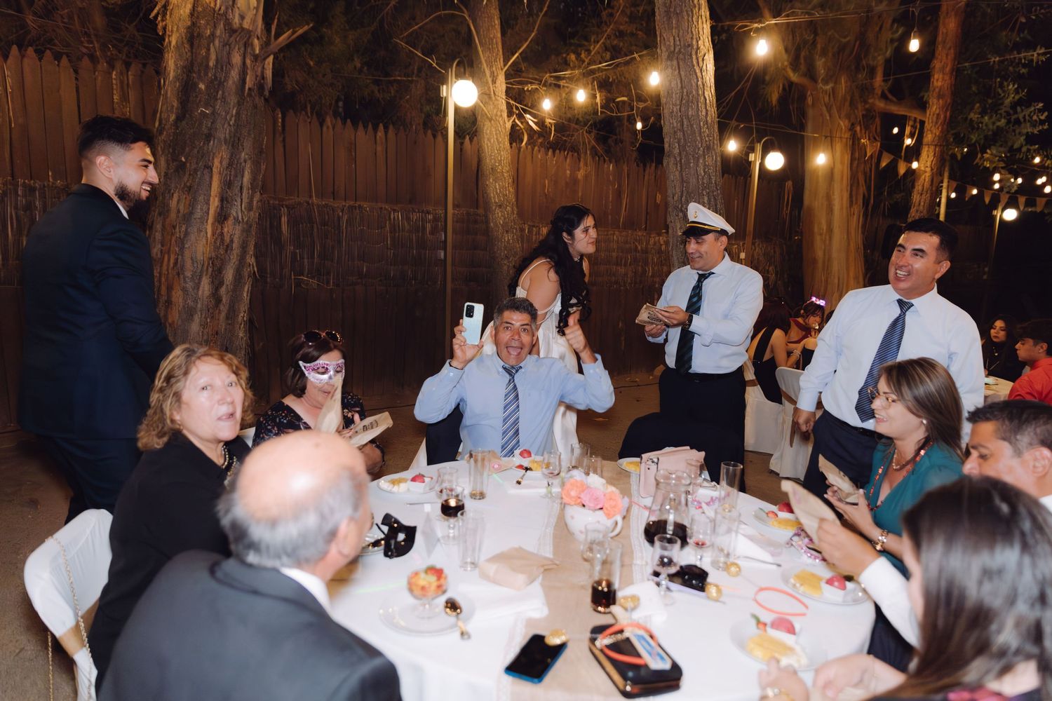 Wedding guests raising phones and celebrating at a lively outdoor reception dinner table.