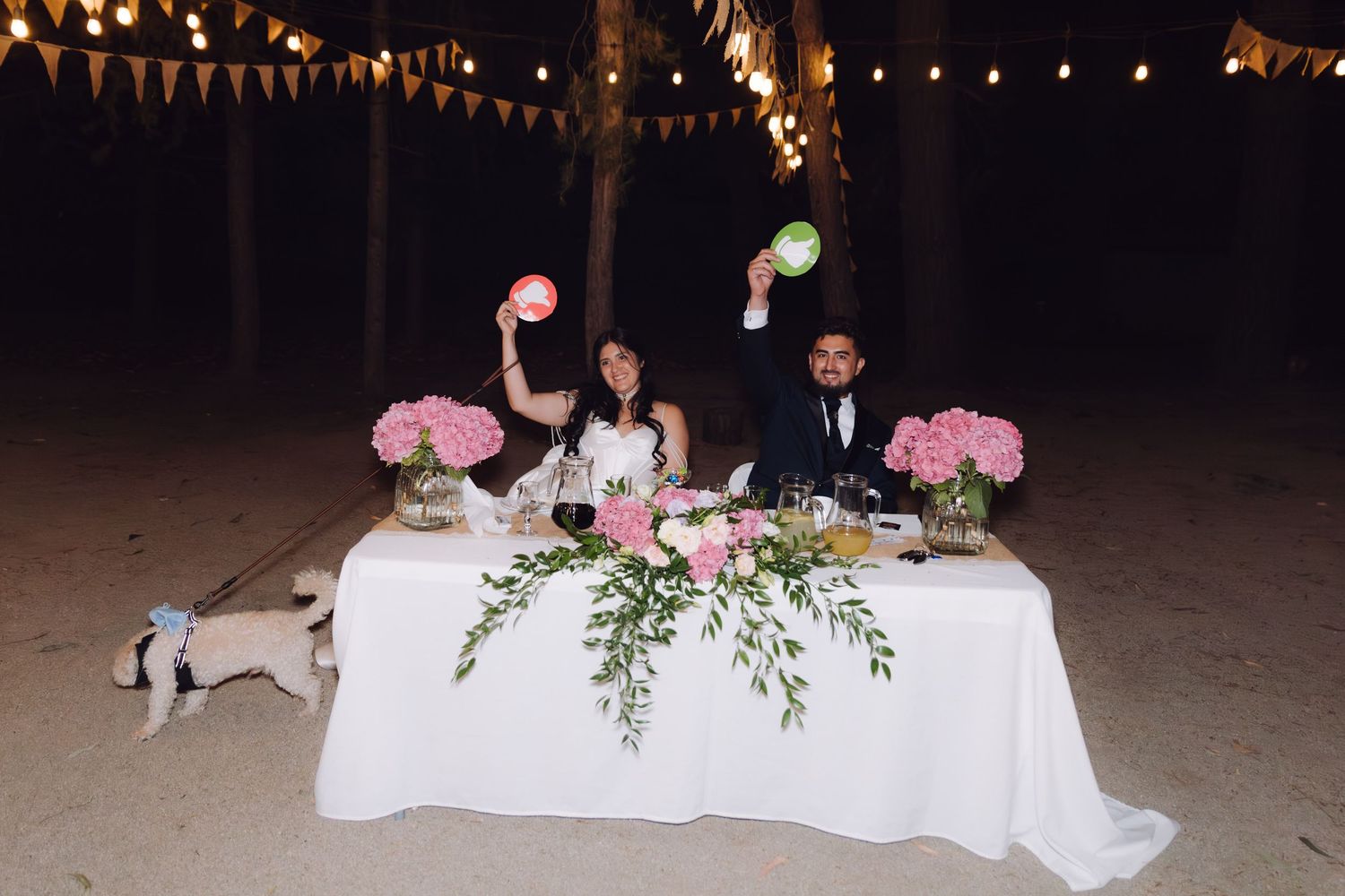 Bride and groom holding colorful signs at a decorated sweetheart table at night.