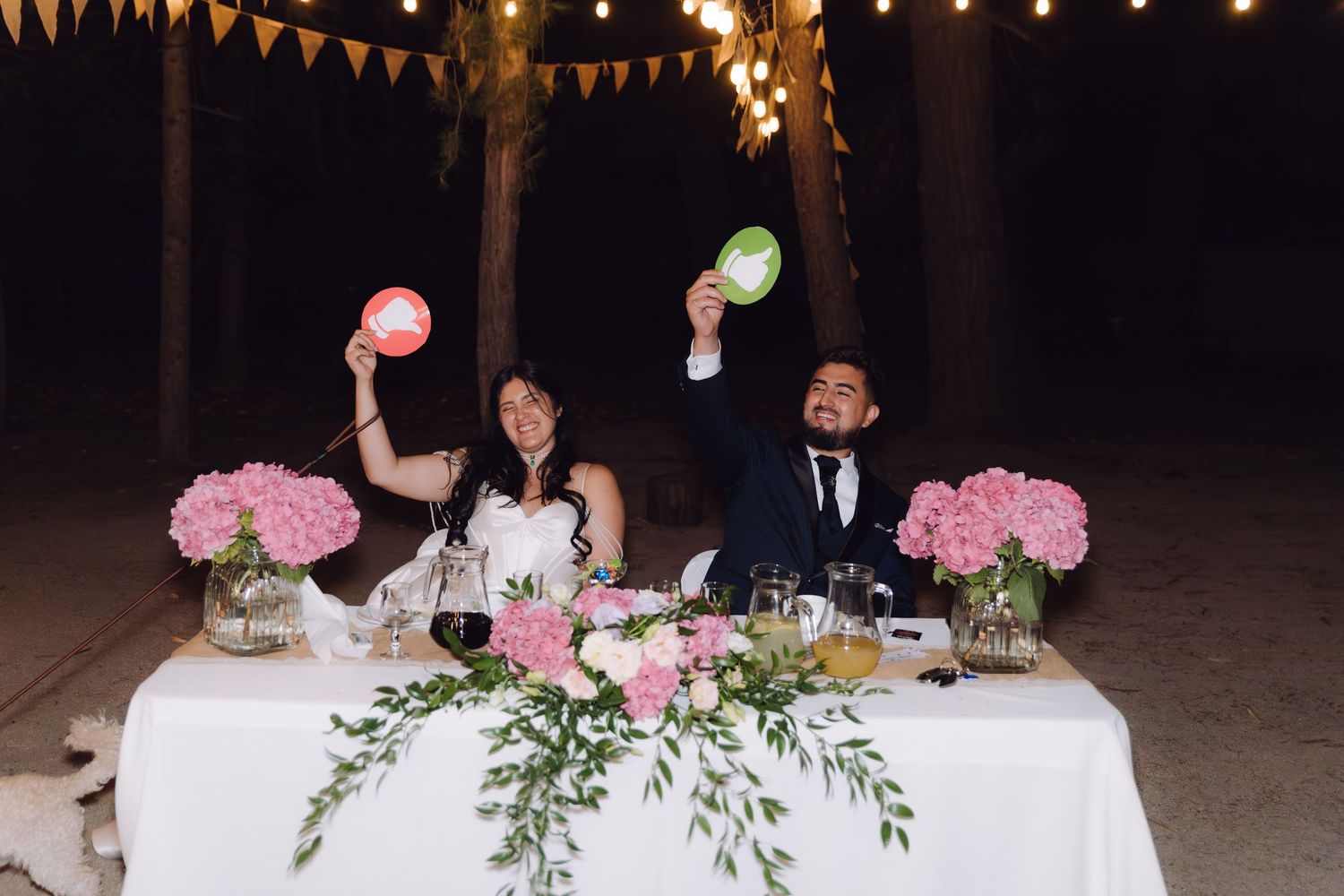Smiling bride and groom holding up signs at a floral-decorated outdoor wedding table.
