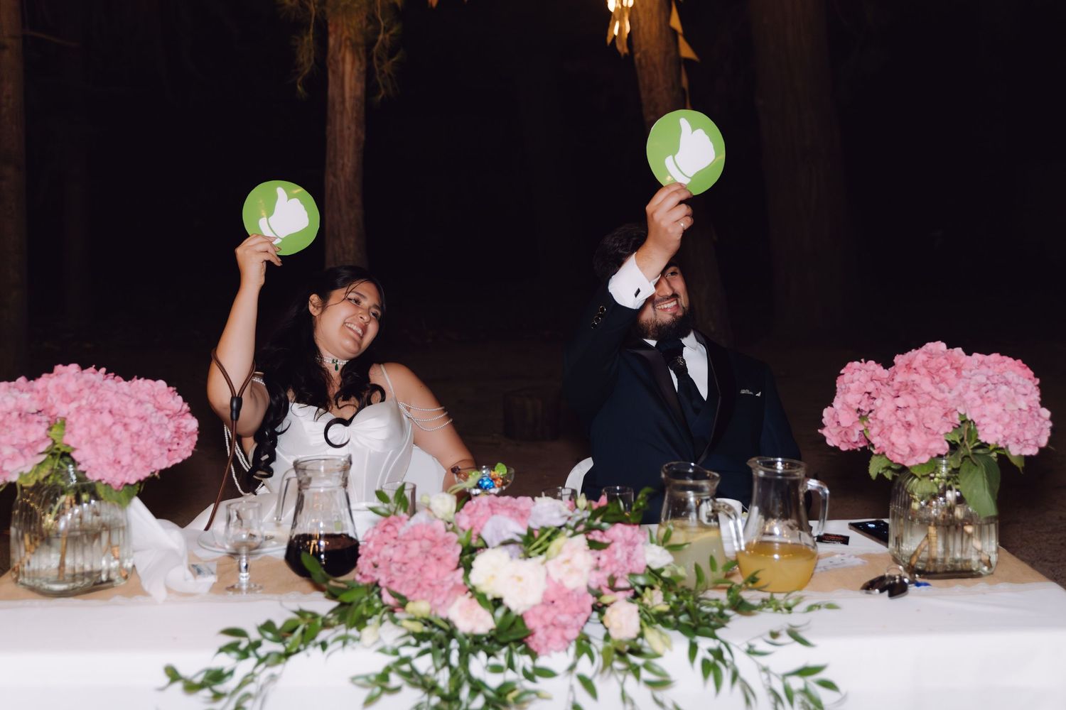 Bride and groom holding green signs at wedding reception table with pink floral centerpieces.