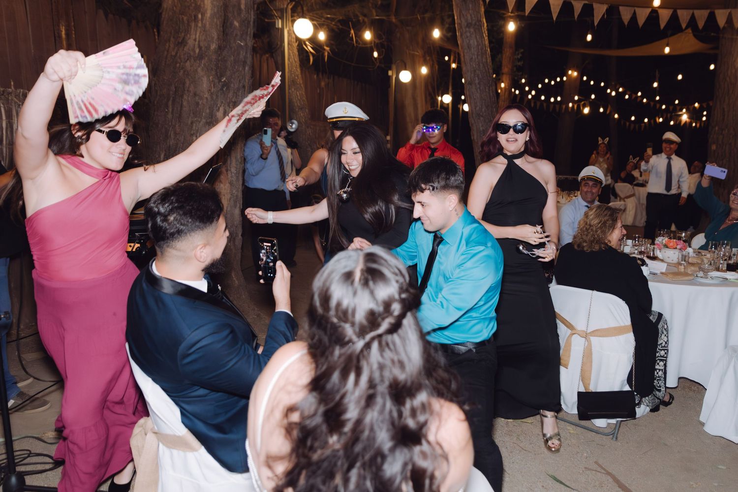Wedding guests dancing energetically at rustic barn reception with string lights overhead.