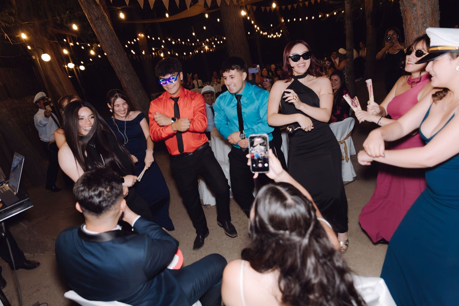Wedding guests dancing and posing for phone camera at festive outdoor evening reception.