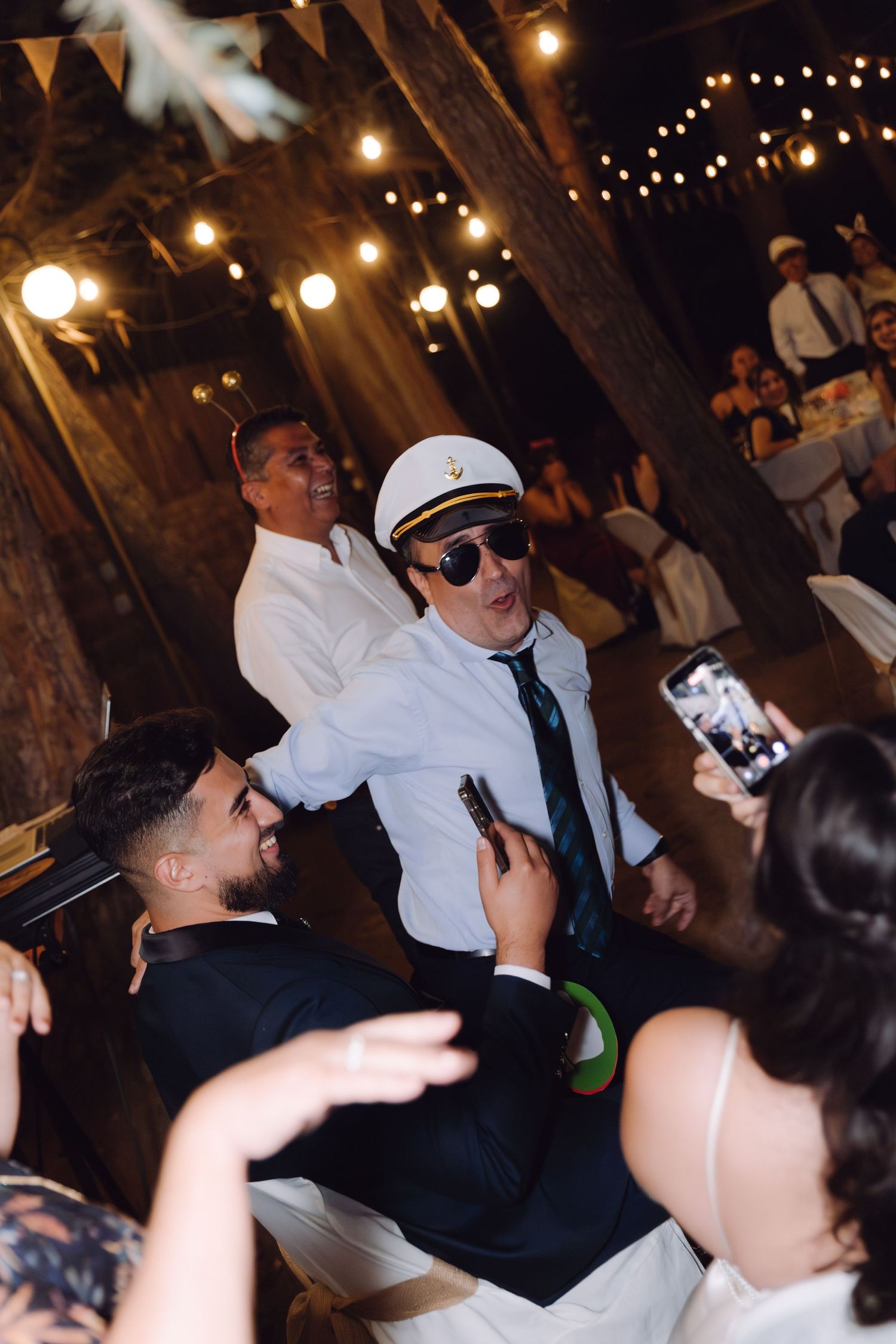 Man in captain hat and sunglasses entertaining bride and groom at wedding reception.