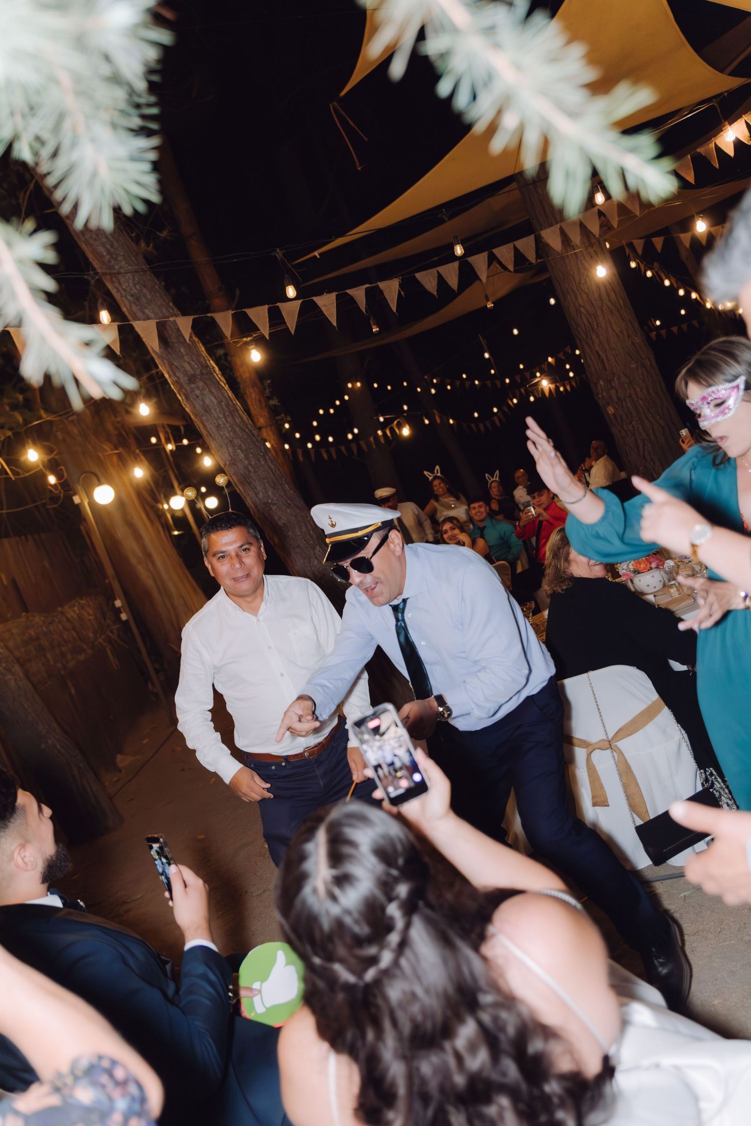 Two men in captain hats dancing at outdoor wedding reception under string lights.