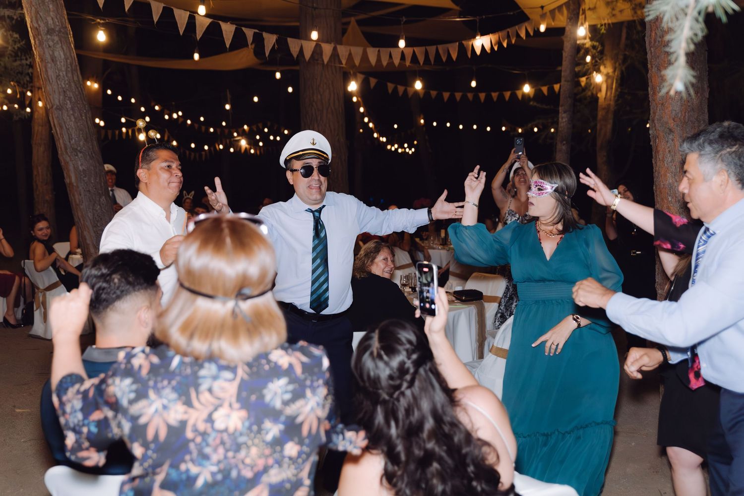 Wedding guests dancing outdoors at night with man in captain hat leading the fun.