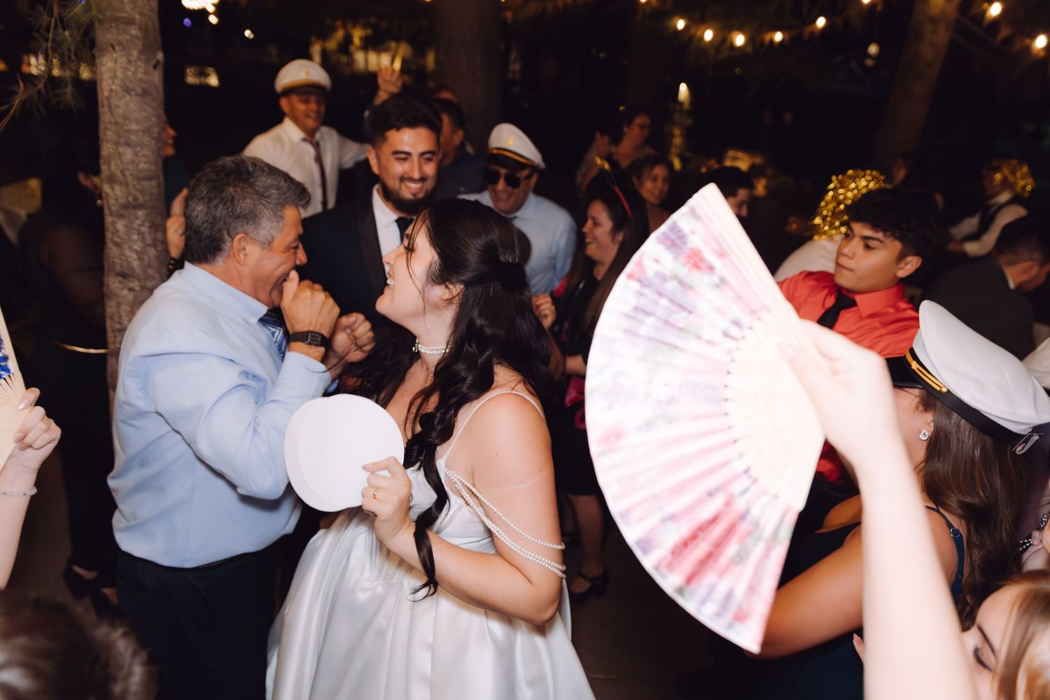 Bride dancing with guests holding decorative fans at lively outdoor wedding reception.