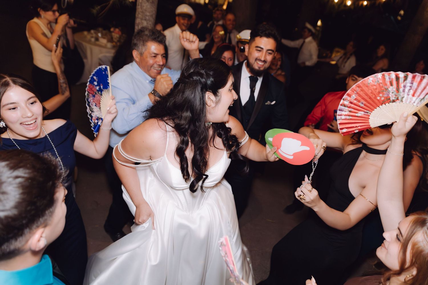 Bride twirling with colorful hand fans surrounded by joyful guests at wedding reception.