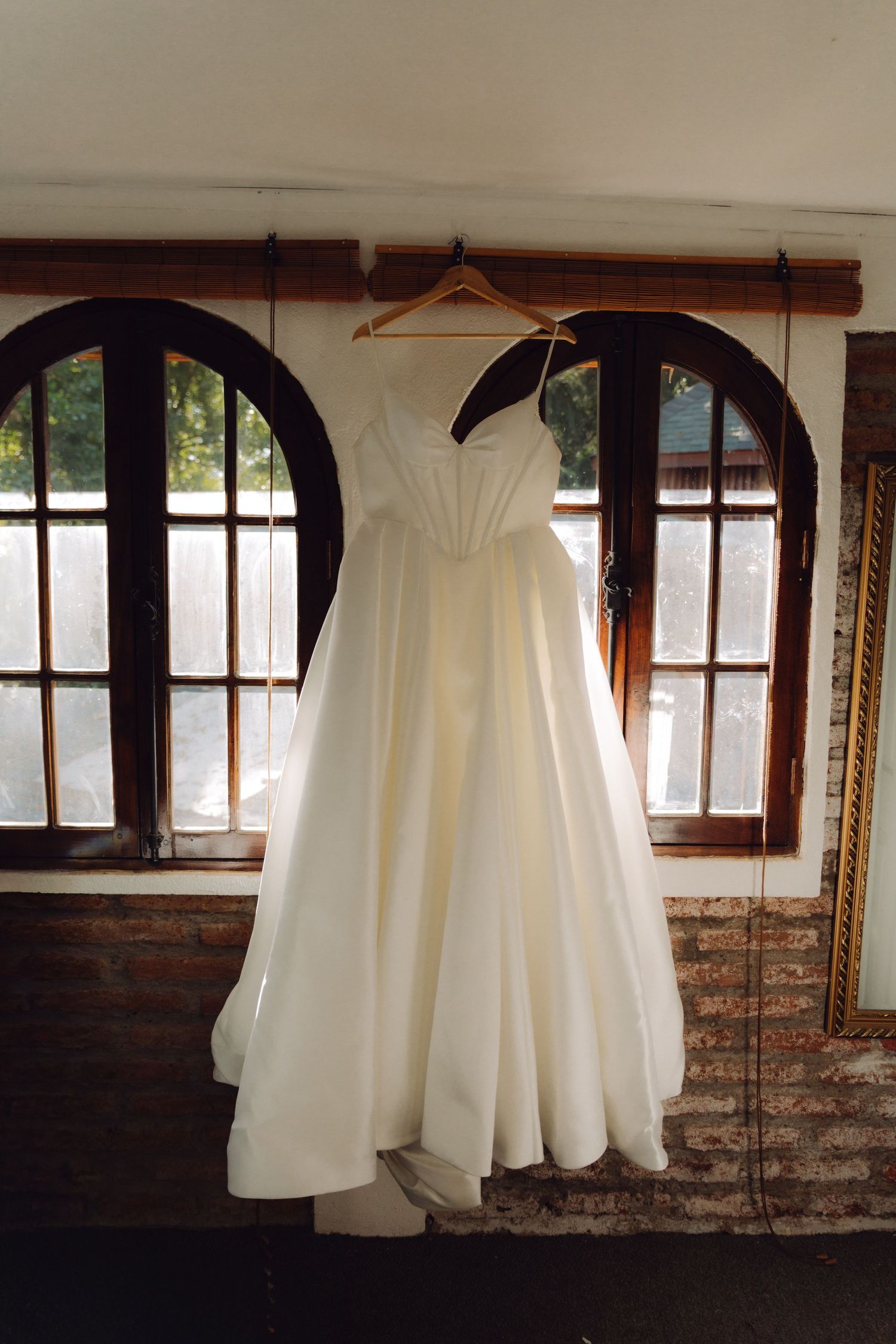 Elegant white A-line wedding dress hanging on wooden hanger in front of arched windows.