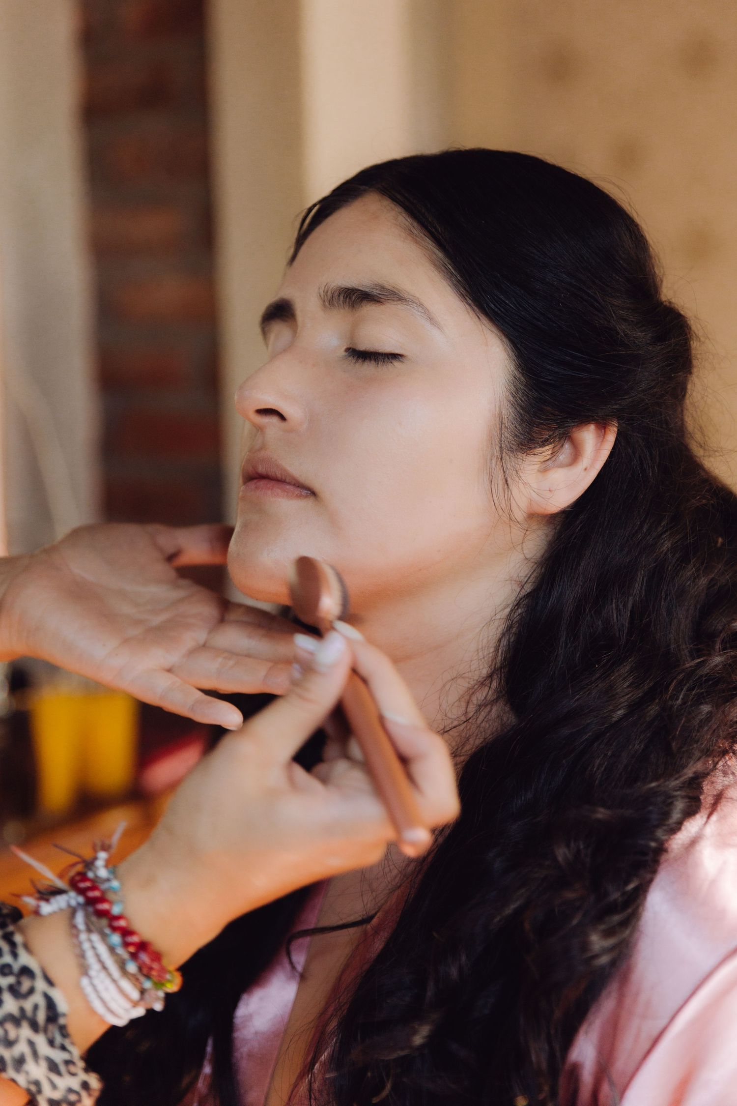 Makeup artist applying foundation to dark-haired bride in pink robe during wedding morning prep.