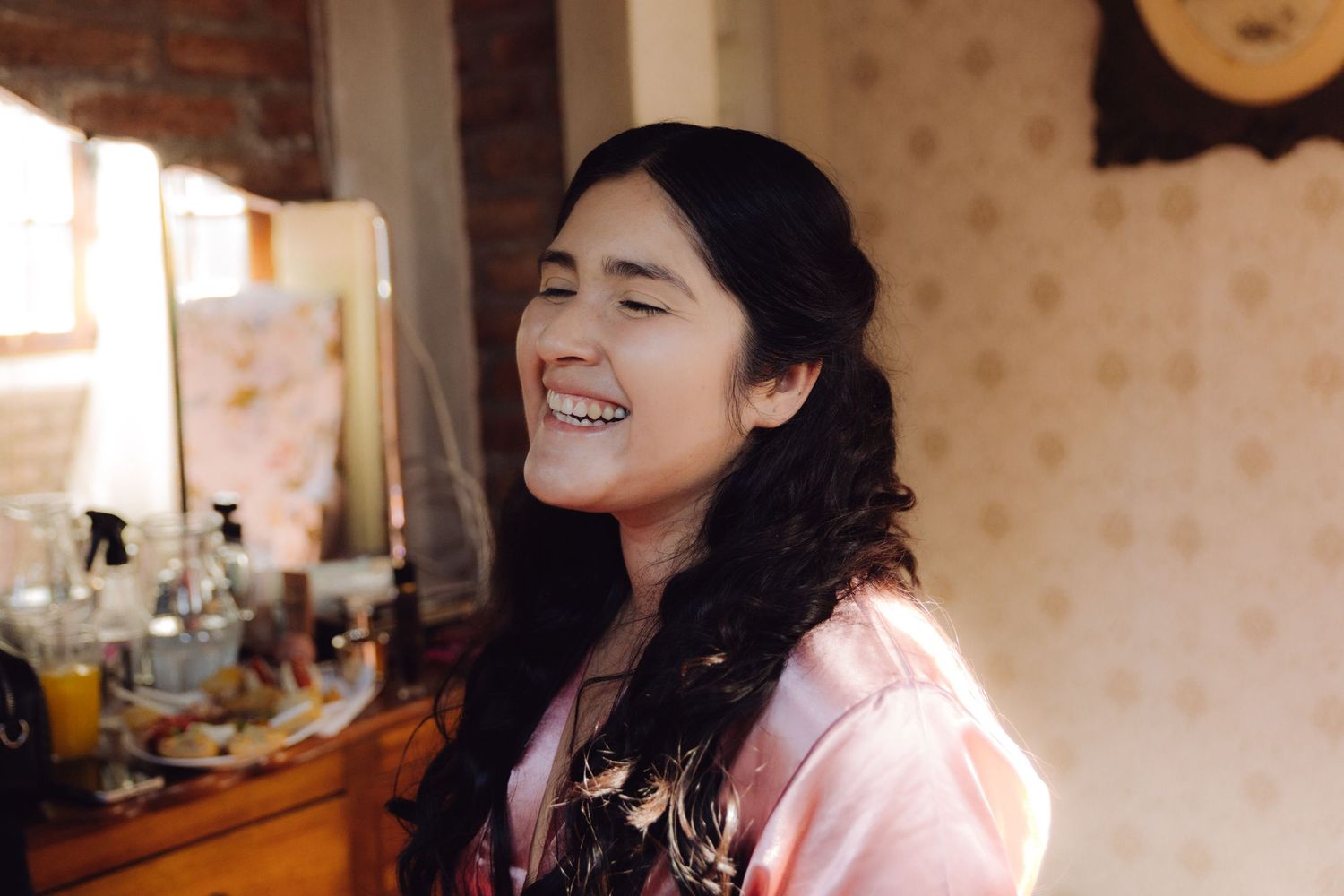 Joyful dark-haired bride in pink robe laughing during wedding morning preparations at vanity.