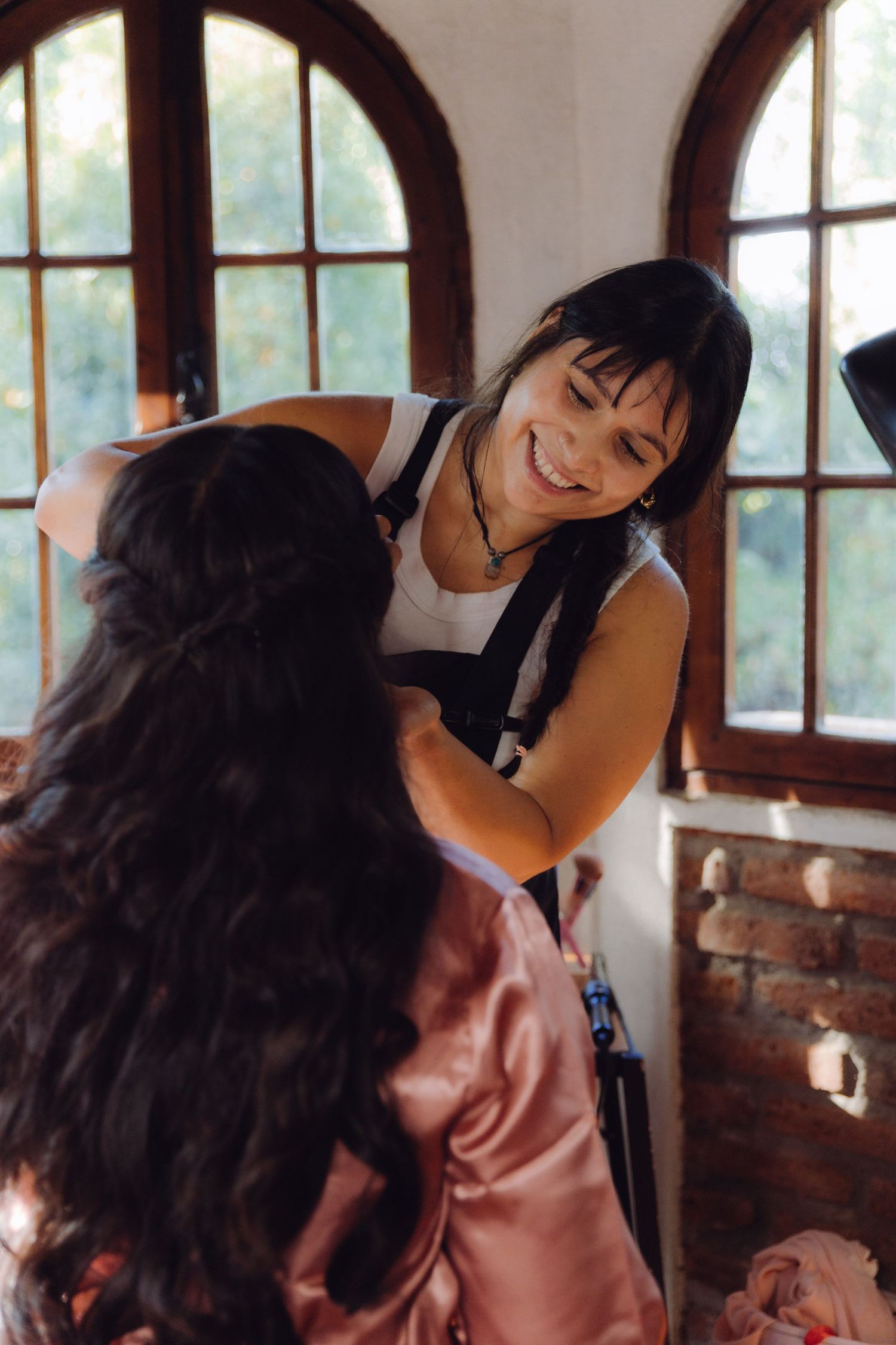 Smiling hairstylist in black apron styling the dark curly hair of a bride in a pink satin robe indoors.