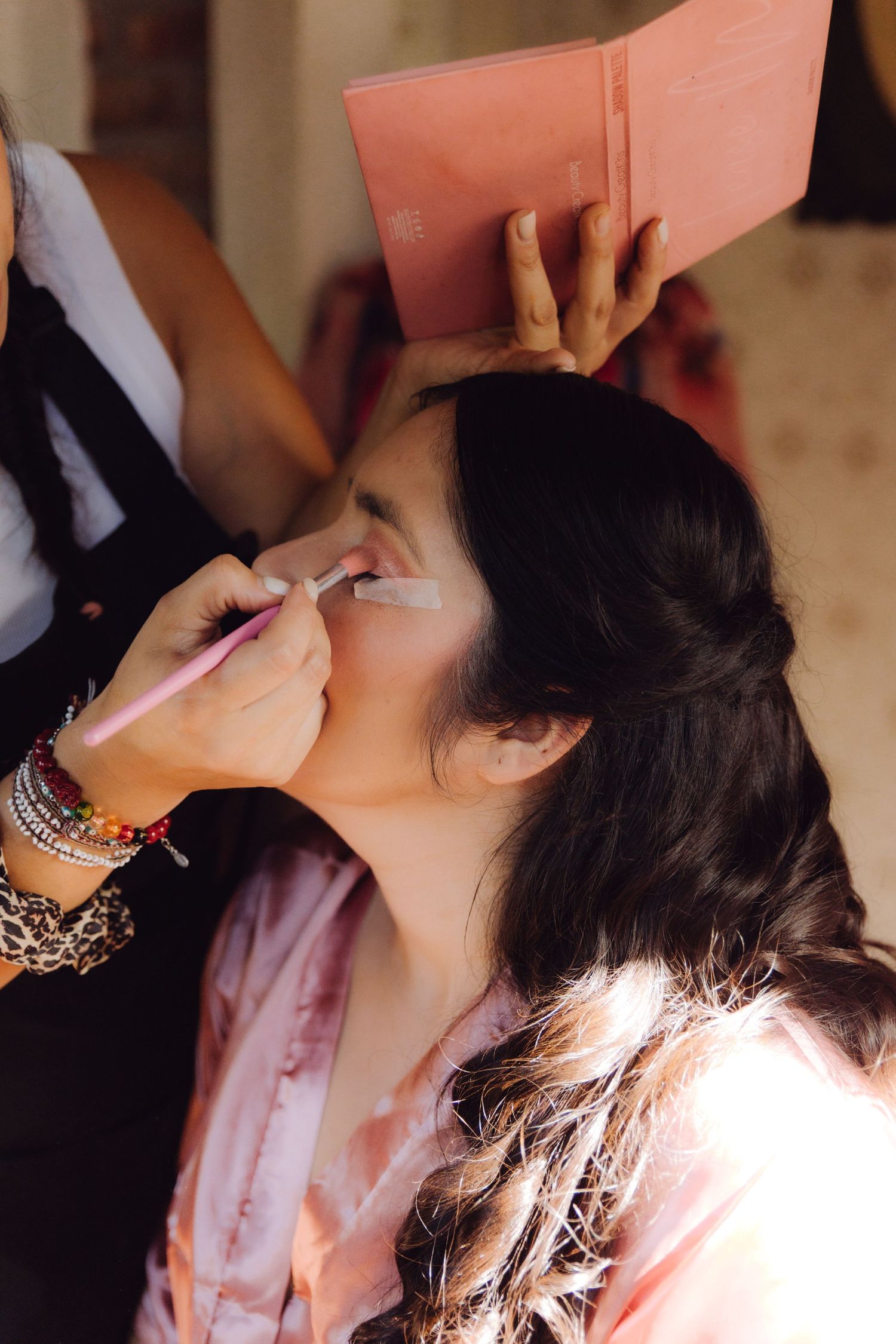Makeup artist applying eyeshadow to a young woman in a pink robe during bridal beauty preparations.