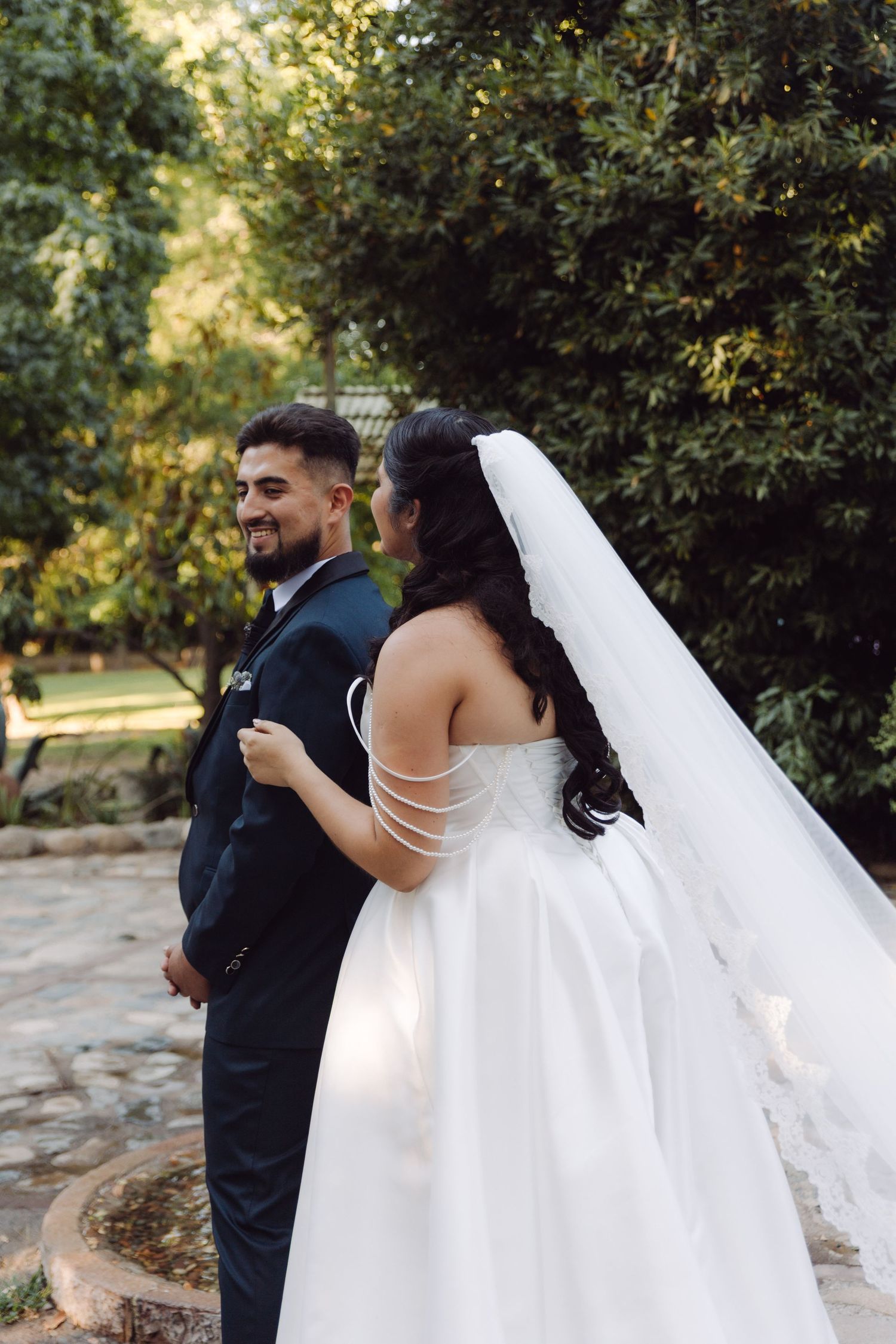Smiling groom in navy suit gazes at bride in strapless white ball gown with long veil in garden setting.