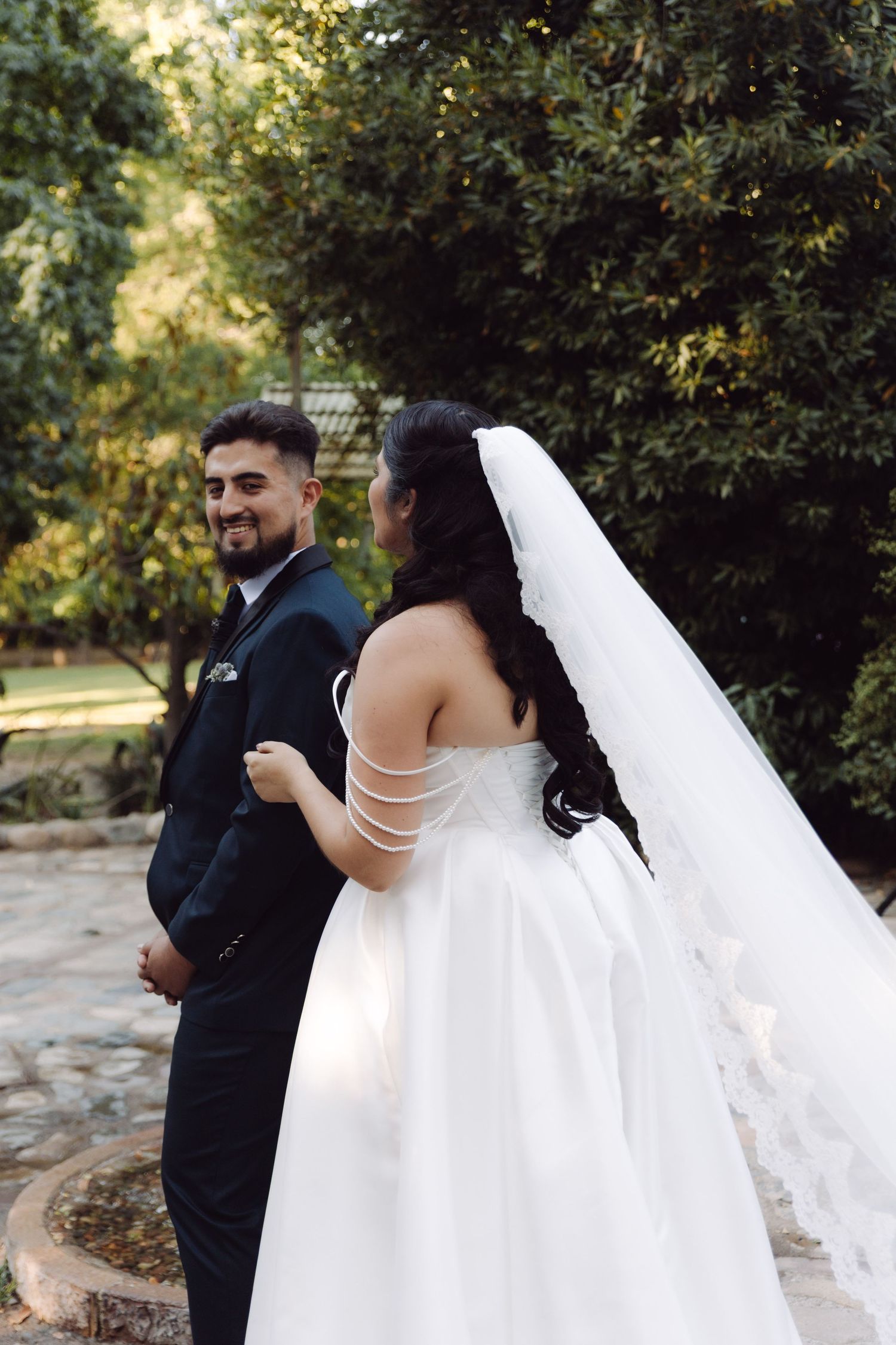 Groom in navy three-piece suit smiles at bride in white strapless gown with long veil among green trees.